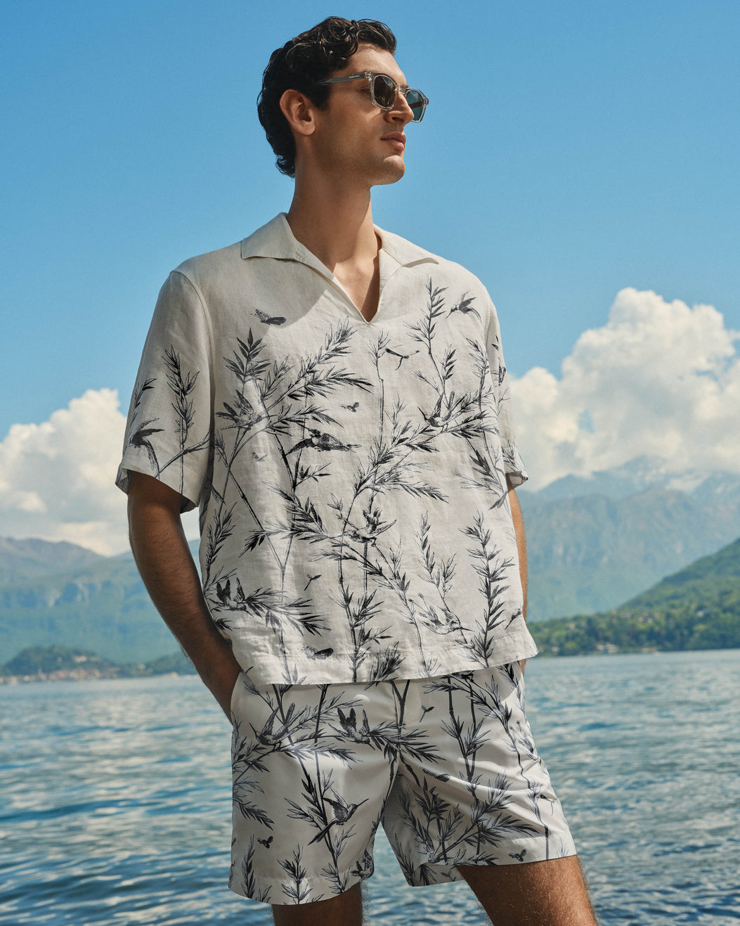 A man wearing a light beige short-sleeve camp collar shirt and matching shorts with a black botanical leaf print, standing outdoors by a lake with mountains and blue sky in the background.