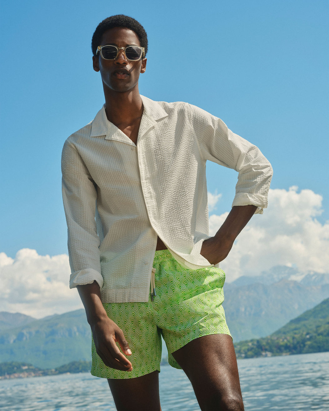 A man wearing a white textured long-sleeve shirt and green patterned swim shorts stands outdoors by a lake with mountains and a blue sky in the background.