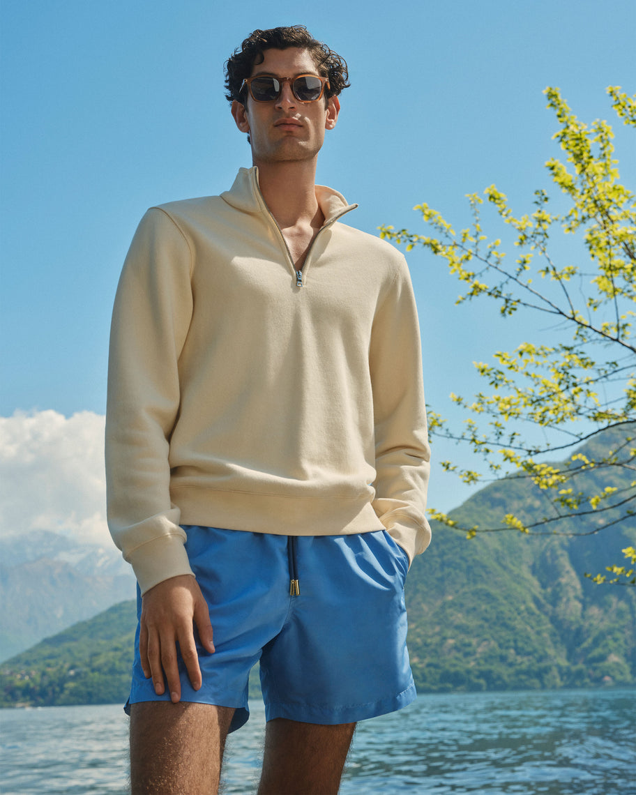 A man wearing a cream quarter-zip pullover and blue swim shorts stands outdoors by a lake with mountains and flowering branches in the background.