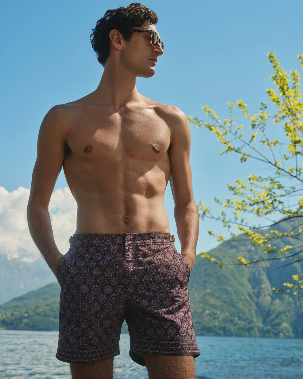 A shirtless man wearing patterned swim shorts and sunglasses stands outdoors by a lake with mountains and blue sky in the background.
