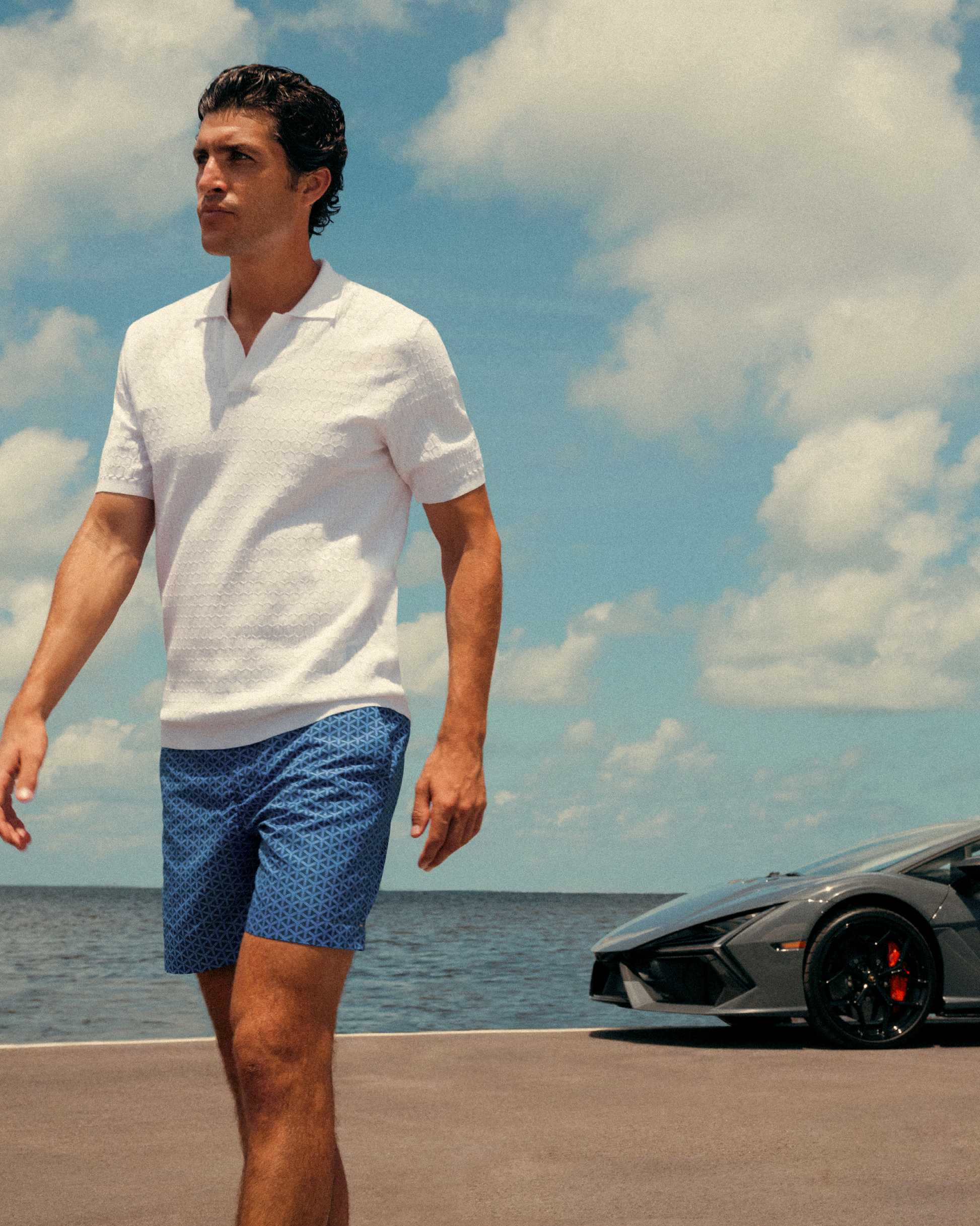Man in a white polo shirt and blue shorts standing by a car with a scenic background