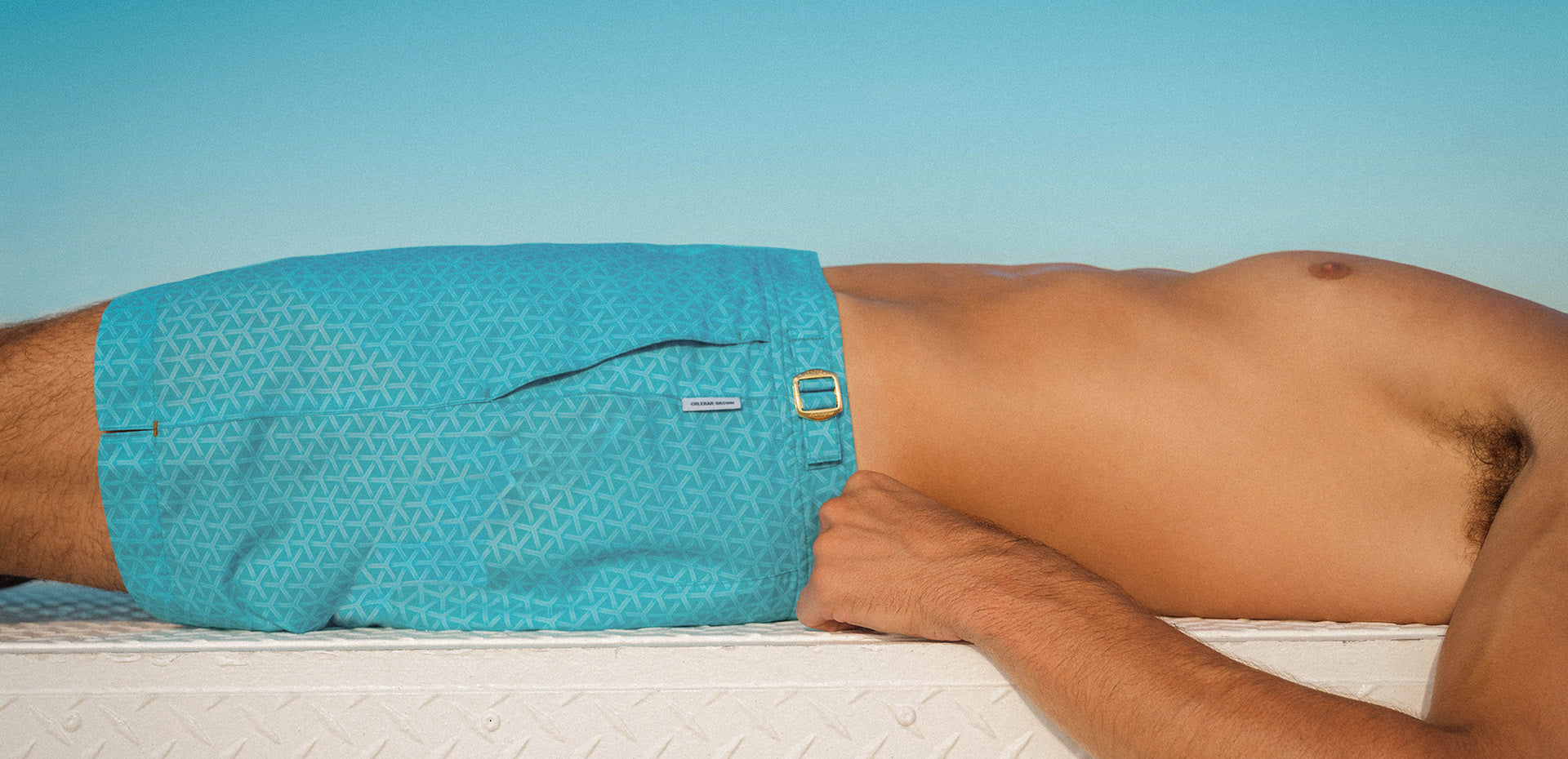 Man lying down wearing turquoise blue swim shorts with side buckle and label branding