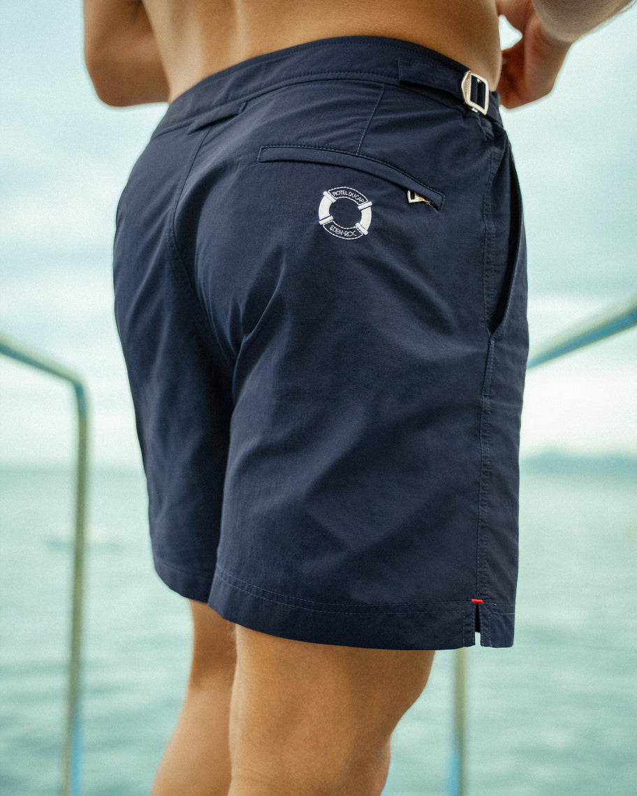 A close-up of a man wearing navy blue board shorts with a white circular logo on the back pocket, photographed outdoors with water and a boat railing in the background.