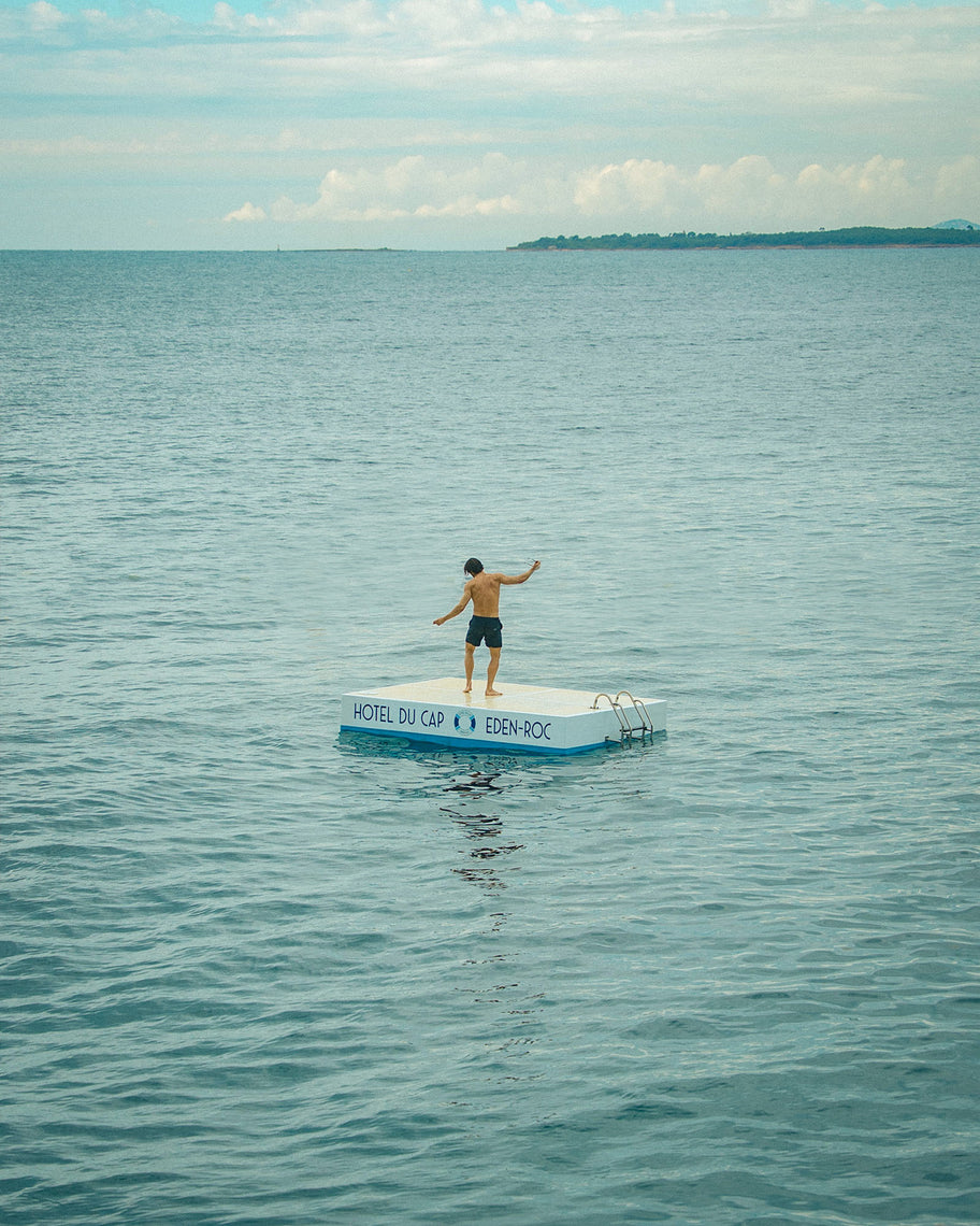 A shirtless man in black swim trunks stands on a small white and blue floating platform in the middle of a calm blue ocean, with distant land and a cloudy sky in the background.