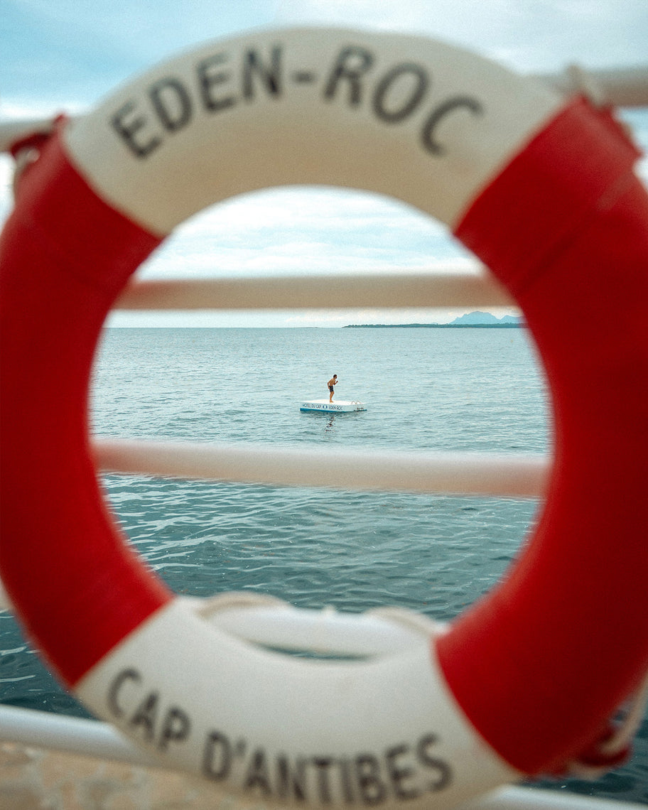 A red and white life preserver ring with the text 'EDEN-ROC' and 'CAP D'ANTIBES' in black lettering, photographed outdoors with a blurred sea and a small paddleboarder visible through the center.