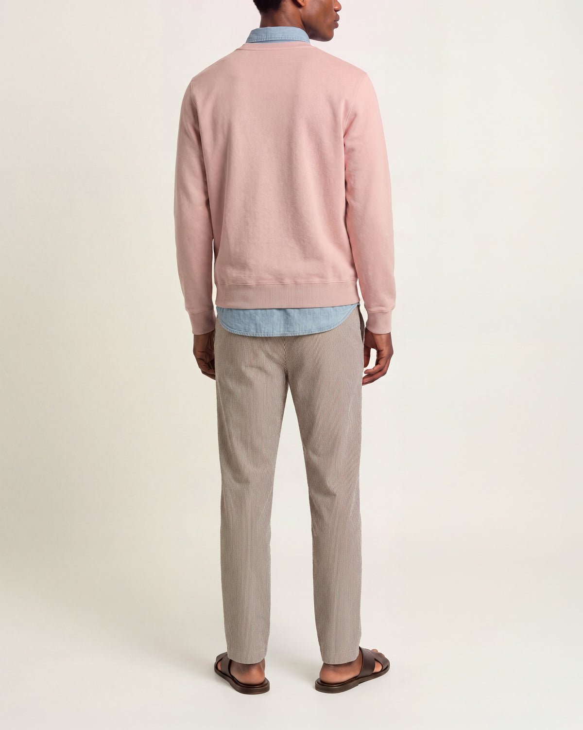 Back view of a man wearing a light pink long sleeved knit sweater over a light blue denim shirt, multicolored trousers, and brown sandals on a plain background