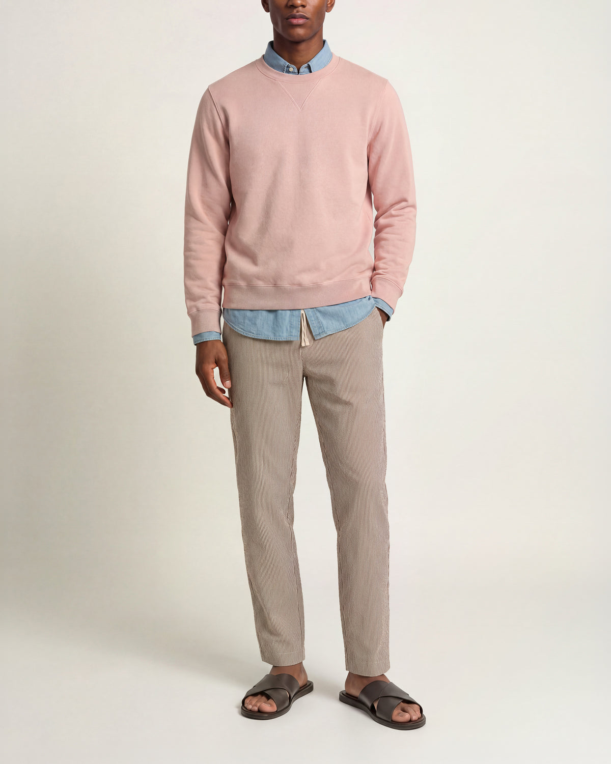 Man wearing a light pink long sleeved knit sweater over a light blue long sleeved denim shirt, multicolored trousers, and brown sandals on a plain background