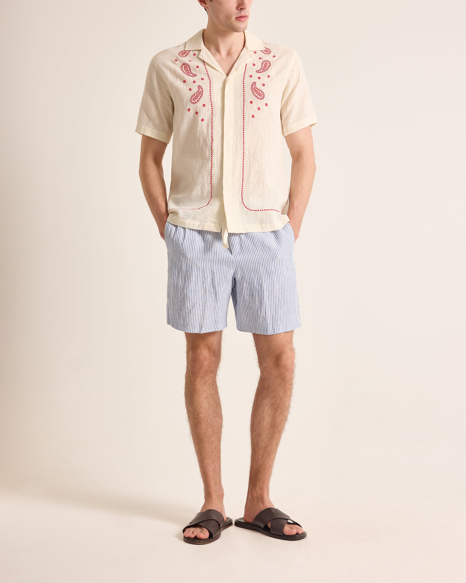 Man wearing a cream embroidered shirt and light blue shorts on a beige background