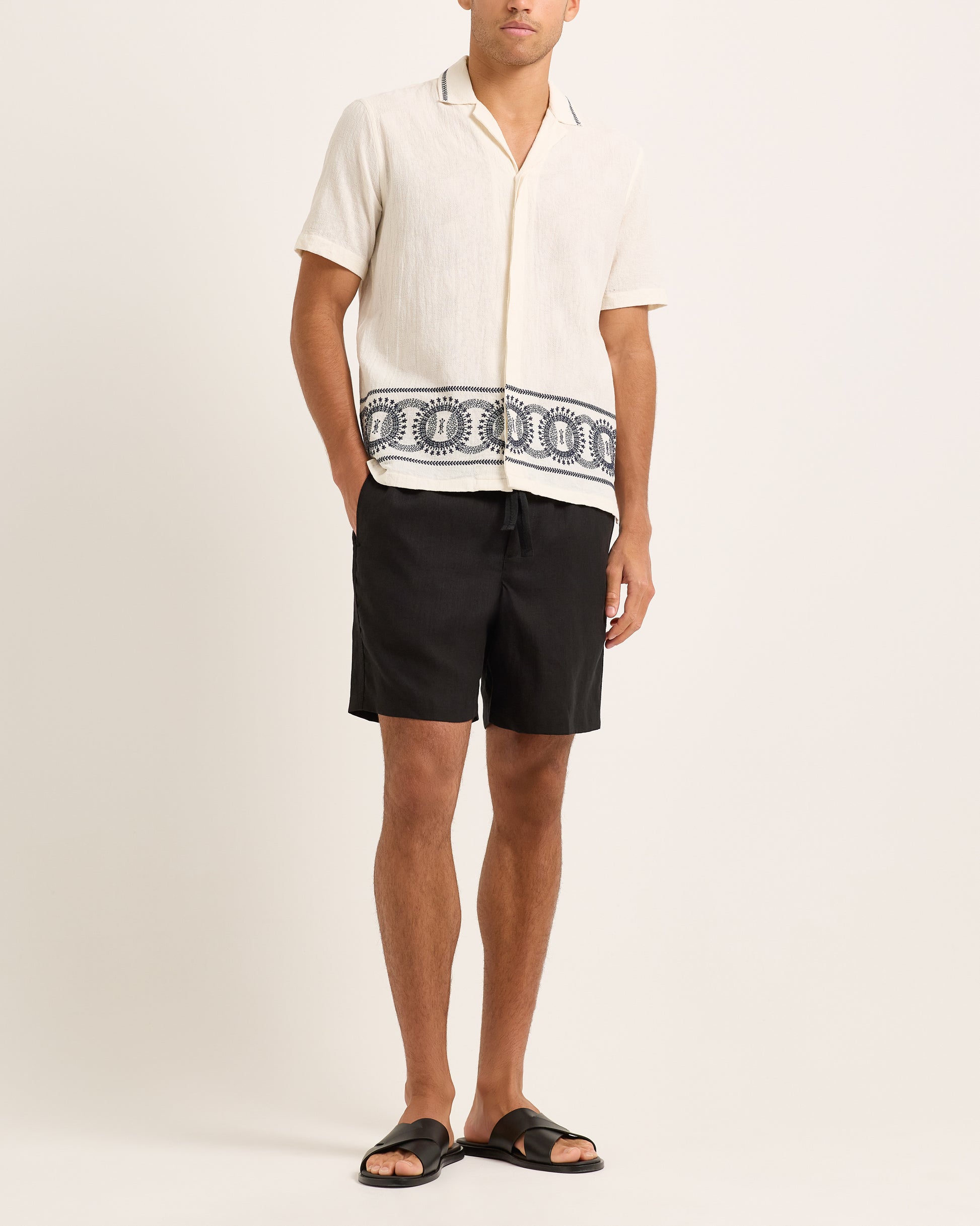 Person wearing a white shirt with a black and white patterned design and black shorts on a beige background