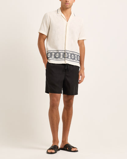 Person wearing a white shirt with a black and white patterned design and black shorts on a beige background
