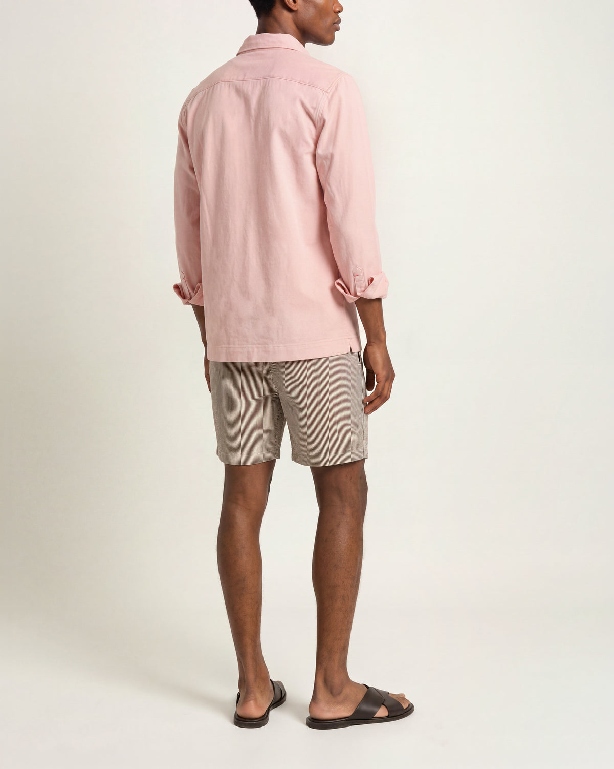 Man viewed from the back wearing light pink long sleeved shirt with multicolored shorts and brown sandals on a plain background