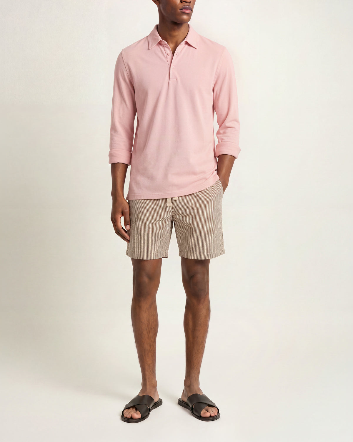 Man wearing light pink long sleeved shirt with multicolored shorts and brown sandals on a plain background