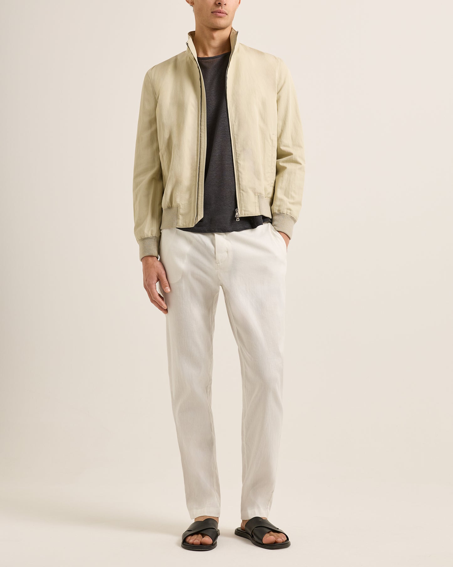 Person wearing a beige bomber jacket, black shirt, white pants, and sandals on a plain background