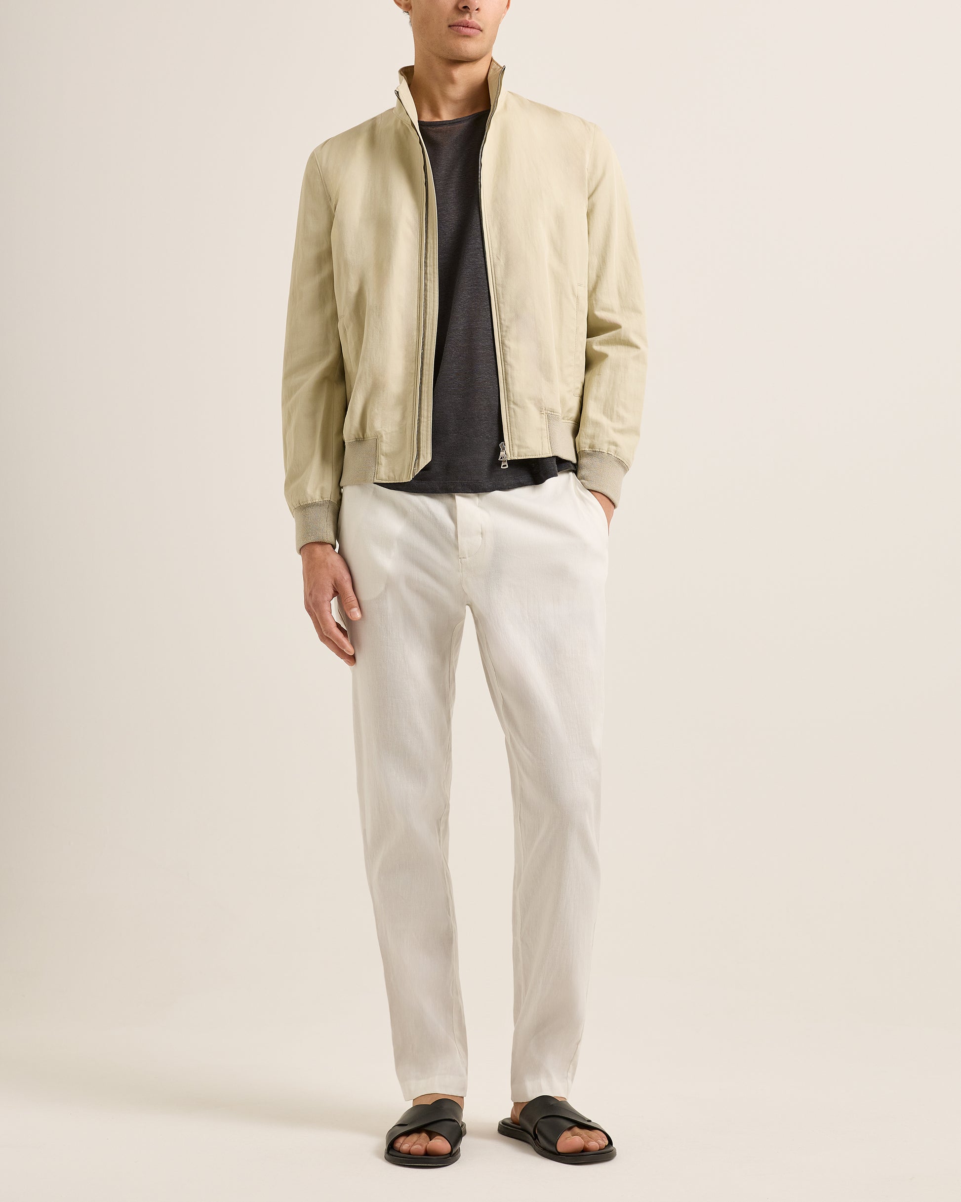 Person wearing a beige bomber jacket, black shirt, white pants, and sandals on a plain background
