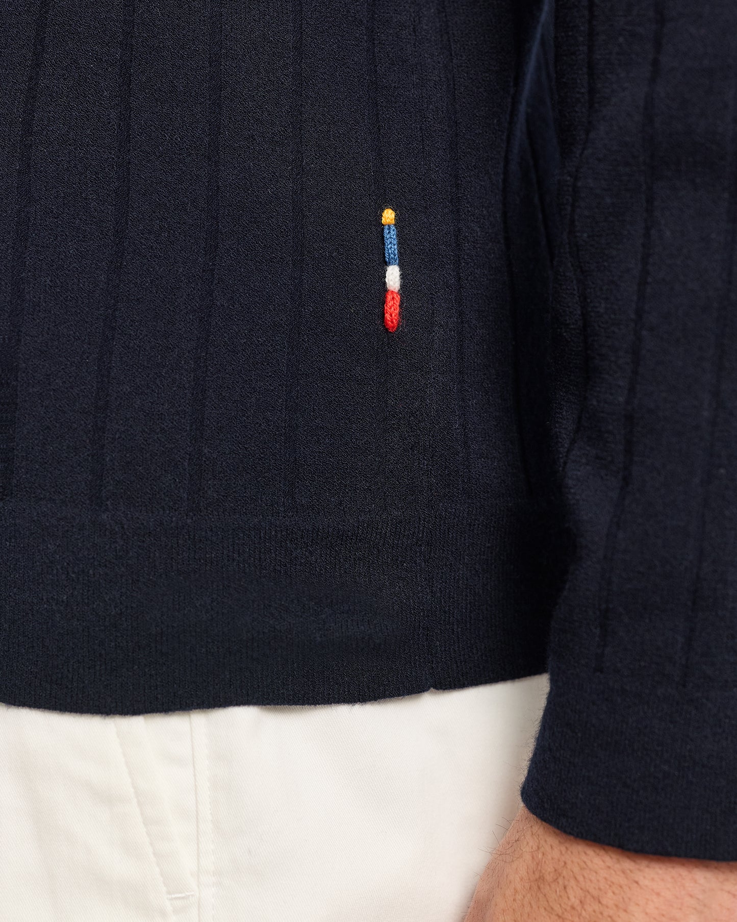 Navy sweater with a colorful embroidered logo worn by a person, with white pants visible.