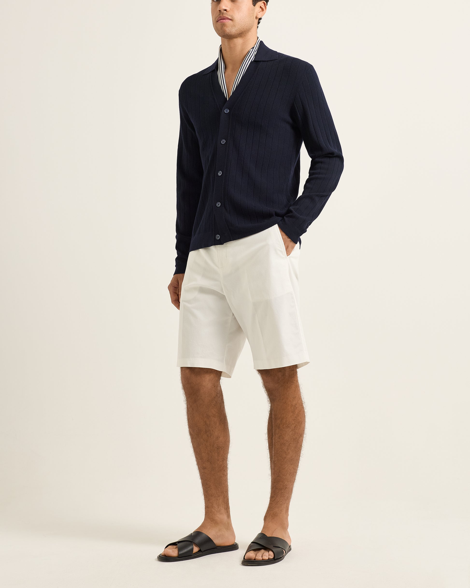 Man wearing a navy cardigan, white shorts, and black sandals on a light beige background