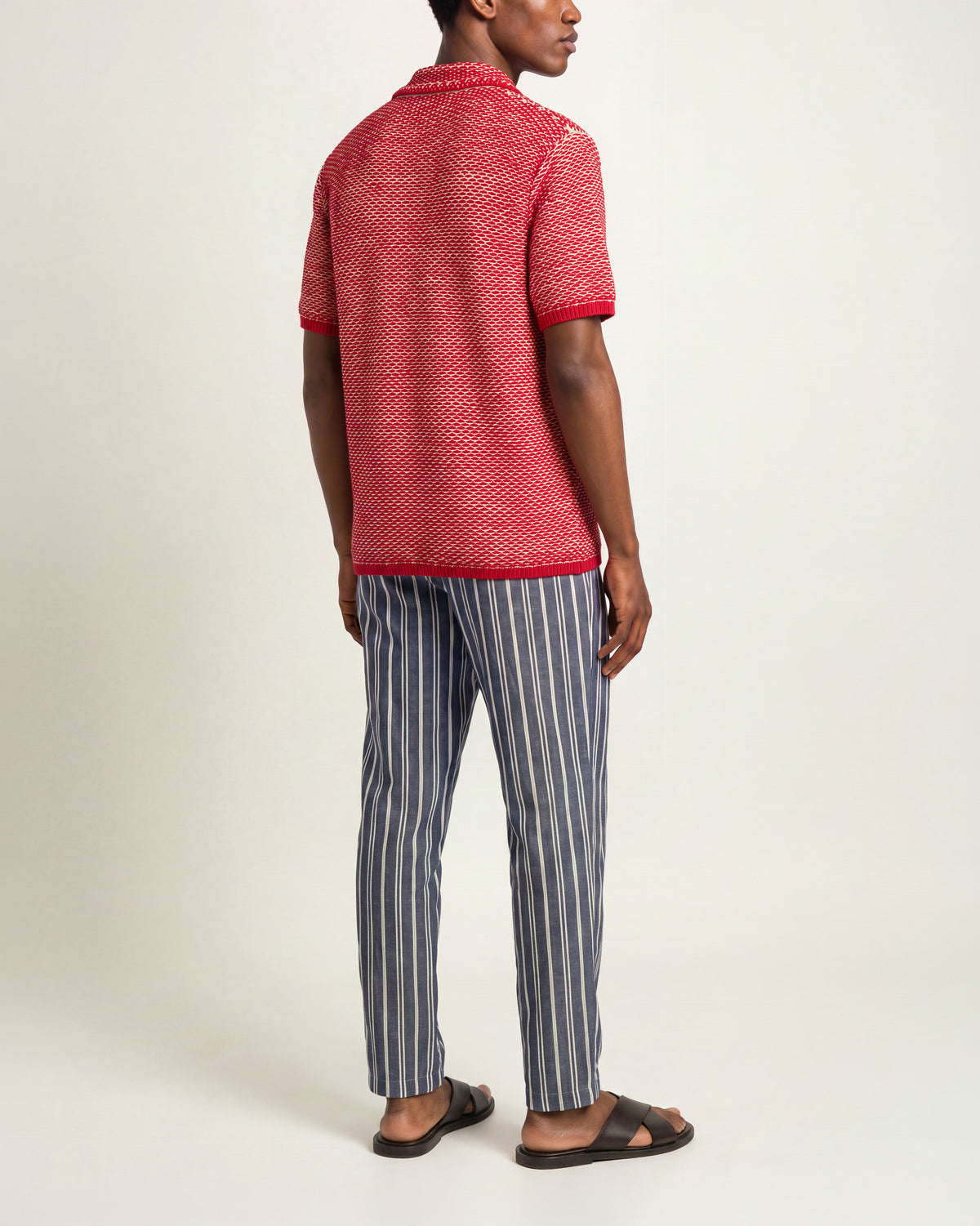 Man wearing red-and-white crochet polo shirt with blue-and-white drawcord waist trousers with brown sandals on a plain background