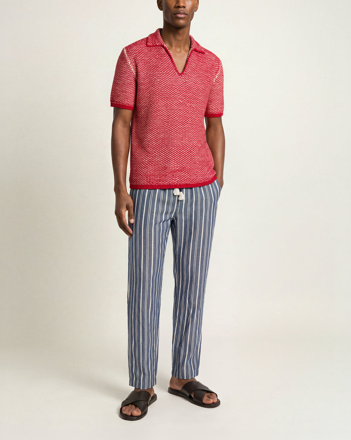 Man wearing red-and-white crochet polo shirt with blue-and-white drawcord waist trousers with brown sandals on a plain background