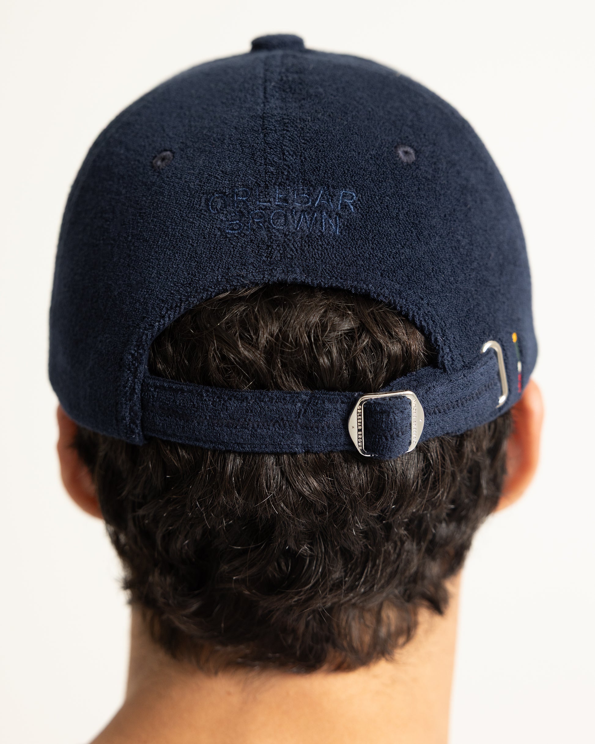 Back view of a person wearing a navy blue baseball cap with 'ORLEBAR BROWN' embroidered on the back, featuring an adjustable strap with a silver metal buckle.