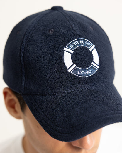 A close-up of a person wearing a dark navy blue baseball cap with a white embroidered life preserver logo featuring the text 'HOTEL DU CAD' and 'EDEN-ROC'.