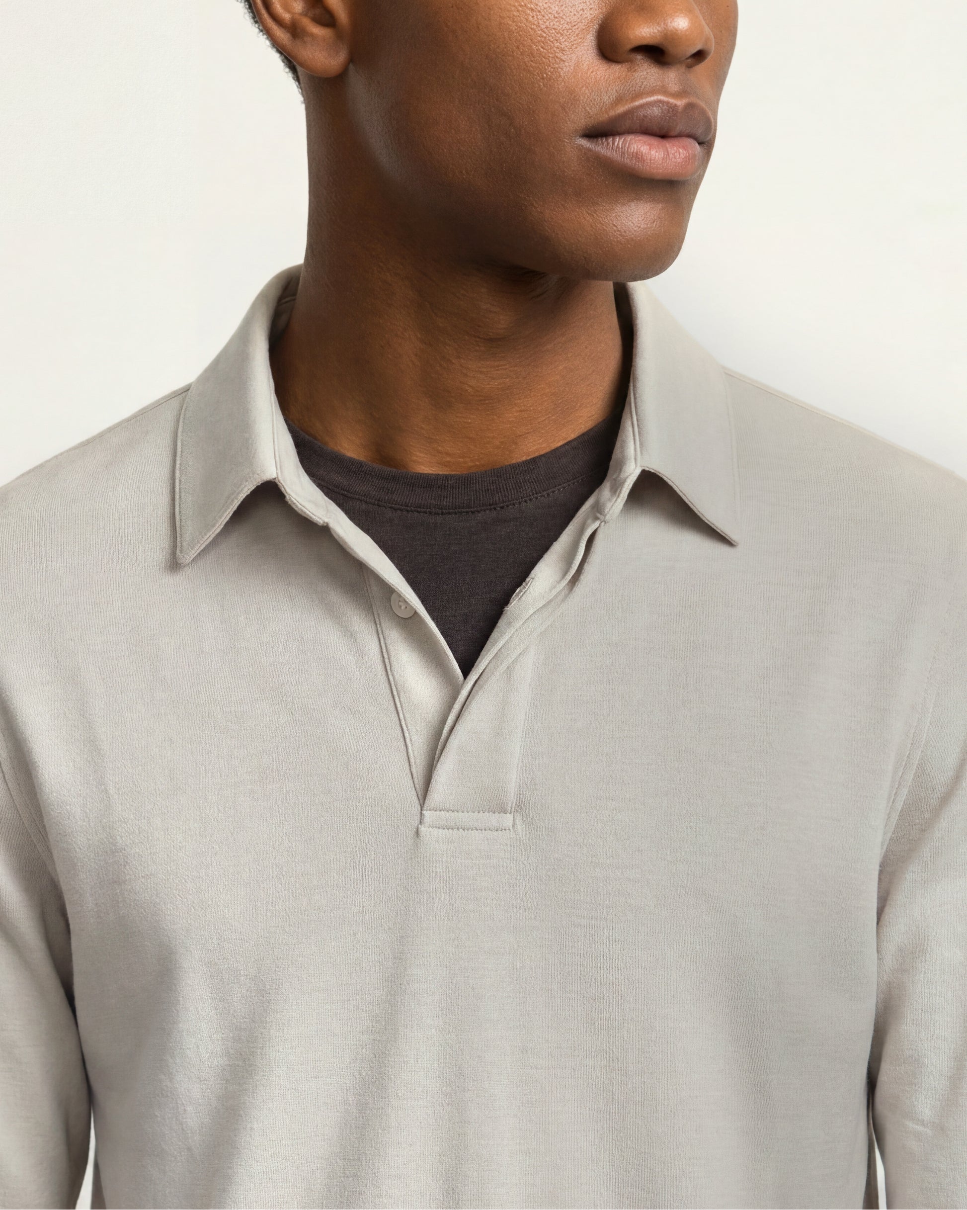 Close-up of a man wearing a light beige long-sleeve polo shirt over a dark charcoal t-shirt, against a plain off-white background.