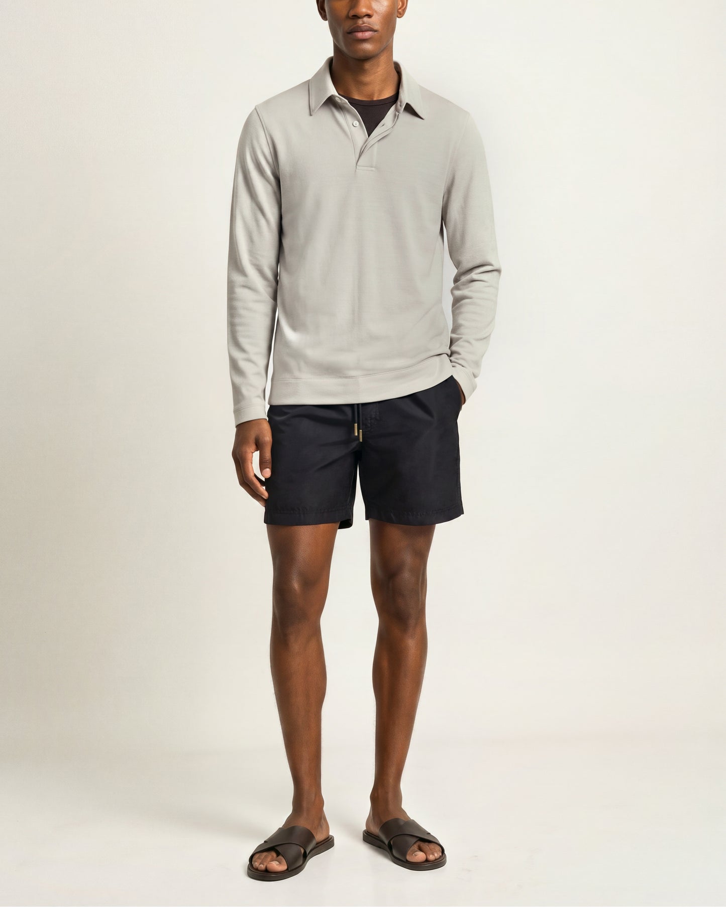 A man modeling a light gray long-sleeve polo shirt with dark navy shorts and dark brown slide sandals, standing against a plain off-white studio background.