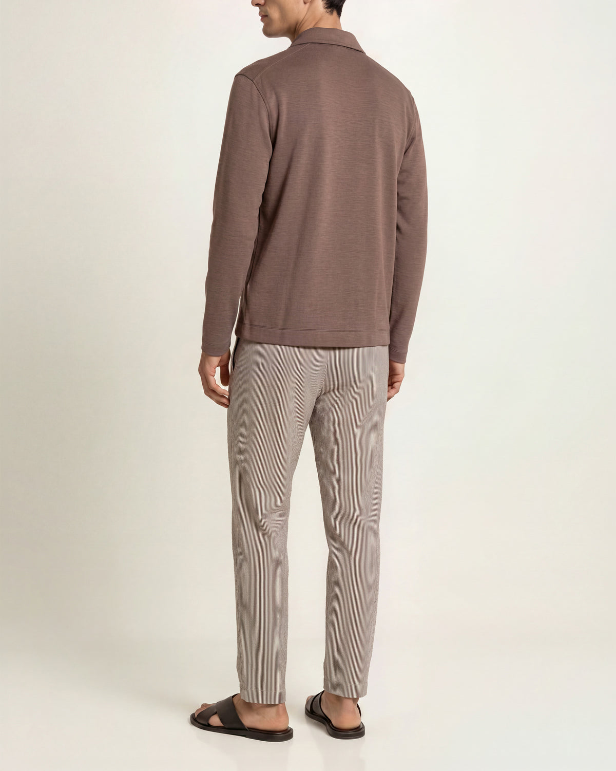 Man wearing brown long sleeve polo shirt with beige t-shirt underneath and brown/white trousers on a plain background
