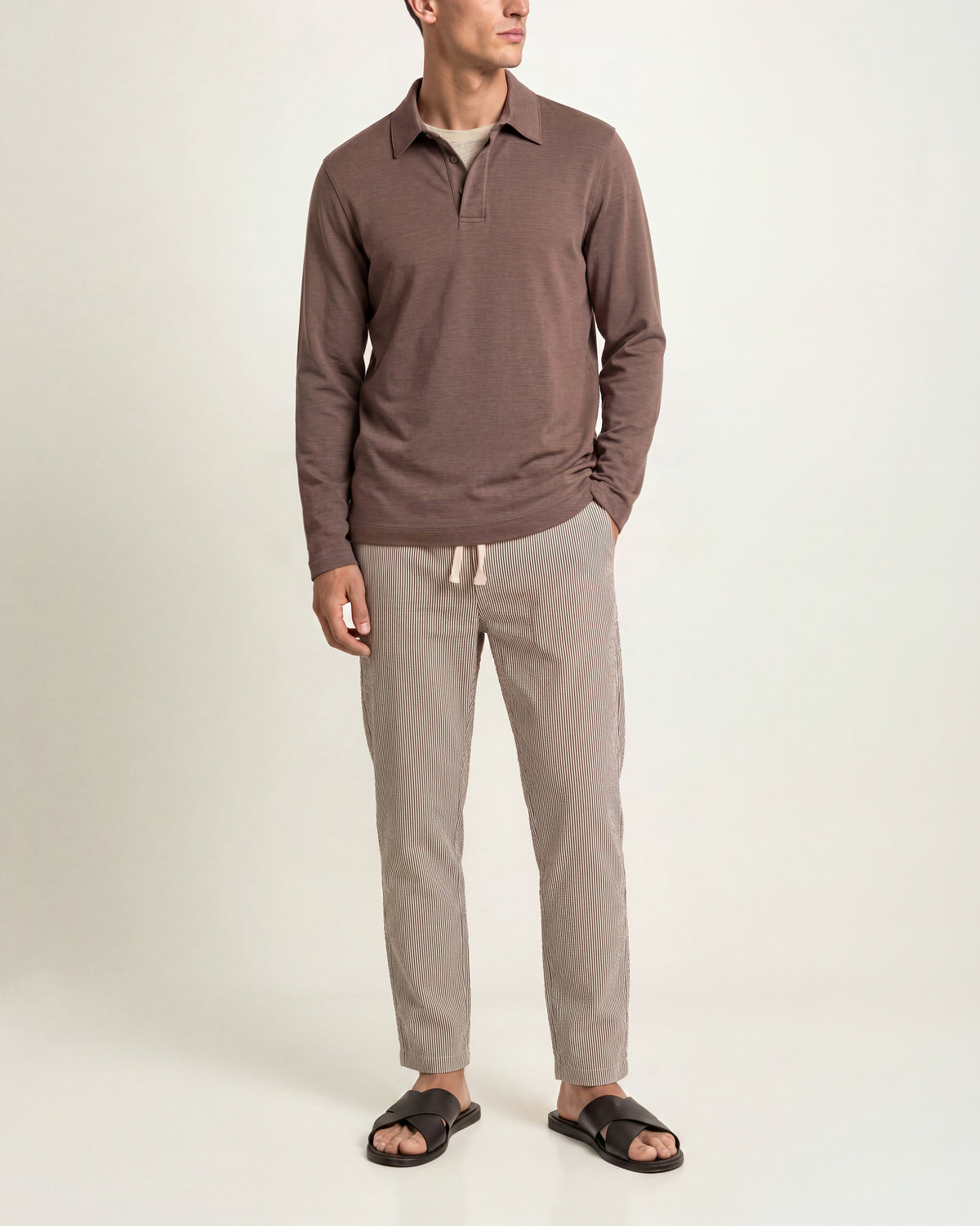 Man wearing brown long sleeve polo shirt with beige t-shirt underneath and brown/white trousers on a plain background