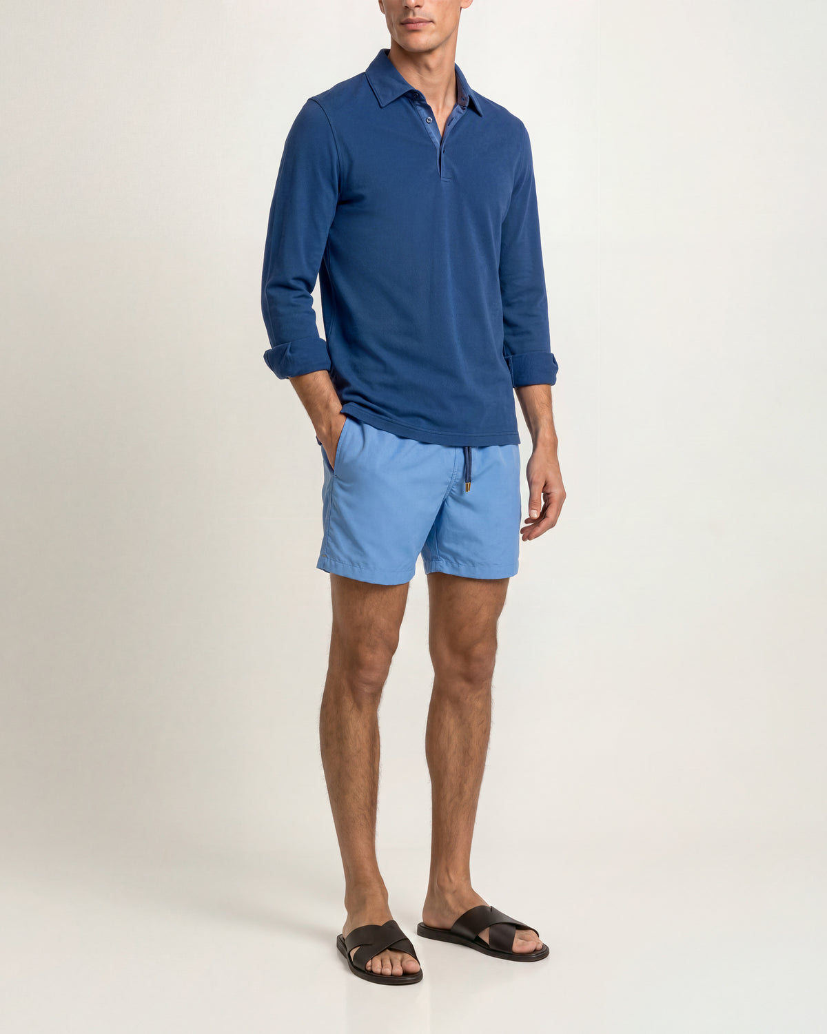 A man modeling a blue long-sleeve polo shirt with light blue swim shorts and black slide sandals, standing against a plain off-white studio background.