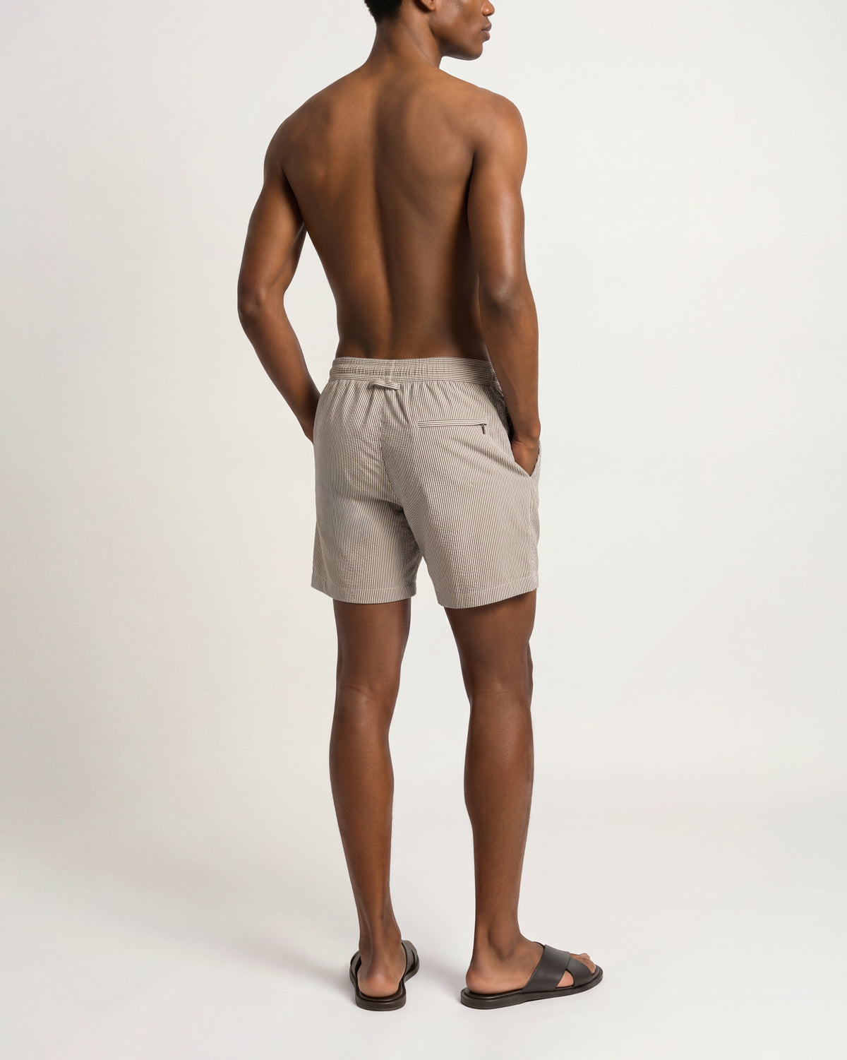 A man shown from the back wearing light beige drawstring shorts and black slide sandals, standing against a plain light gray studio background.