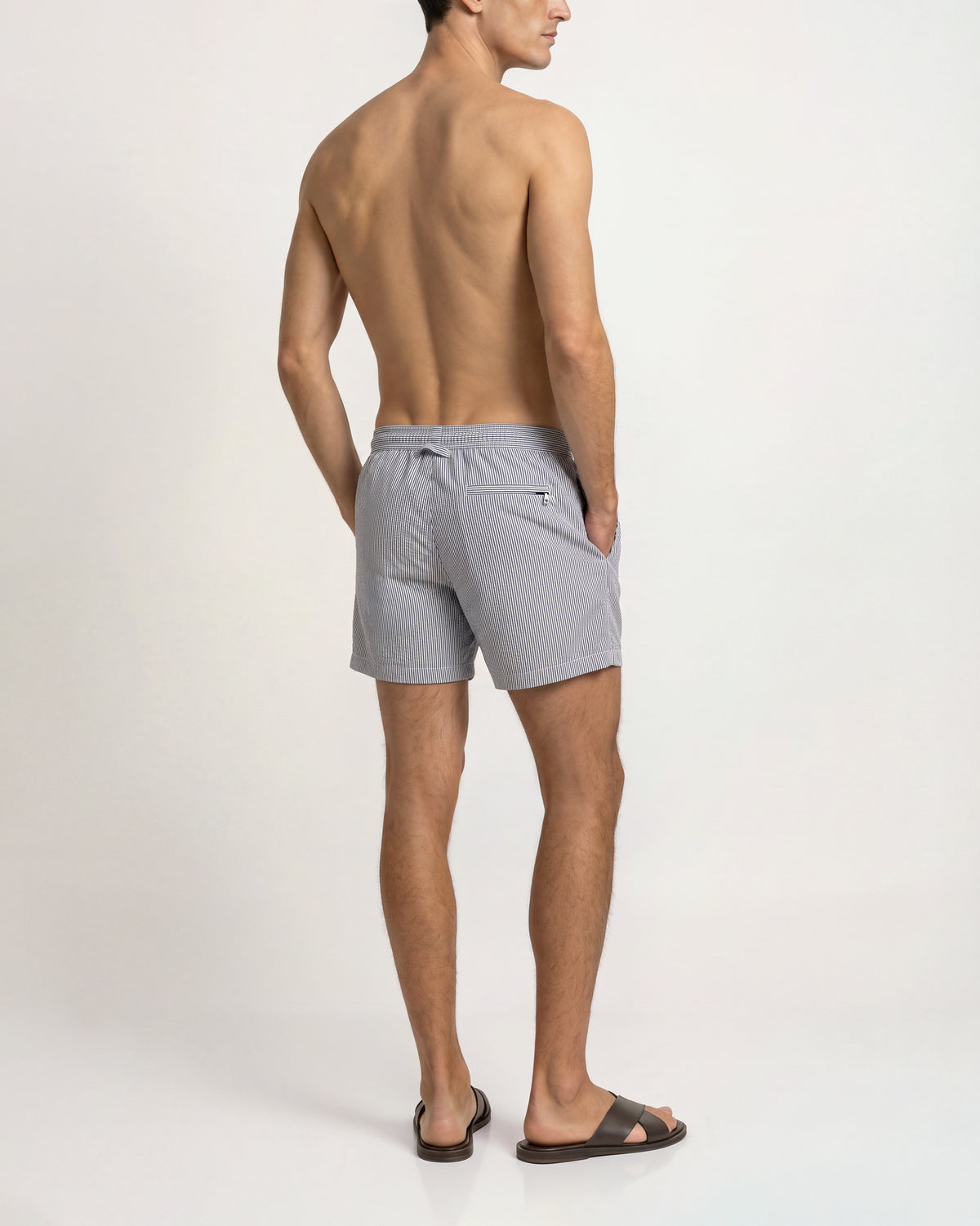 A man shown from the back wearing light gray drawstring swim shorts and dark brown slide sandals against a plain white studio background.