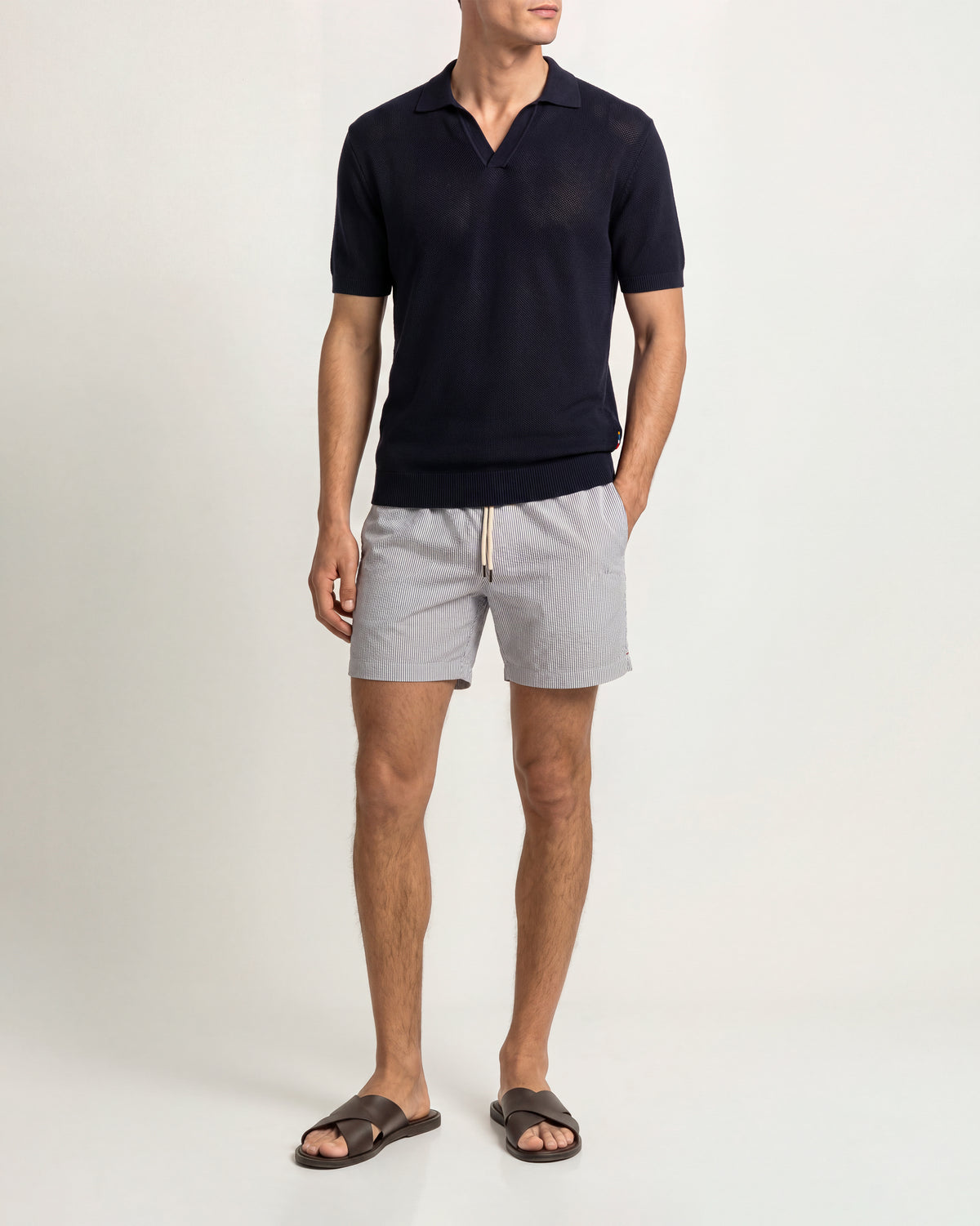 A man modeling a dark navy short-sleeve knit polo shirt paired with light gray drawstring shorts and dark brown slide sandals, photographed against a clean off-white studio background.