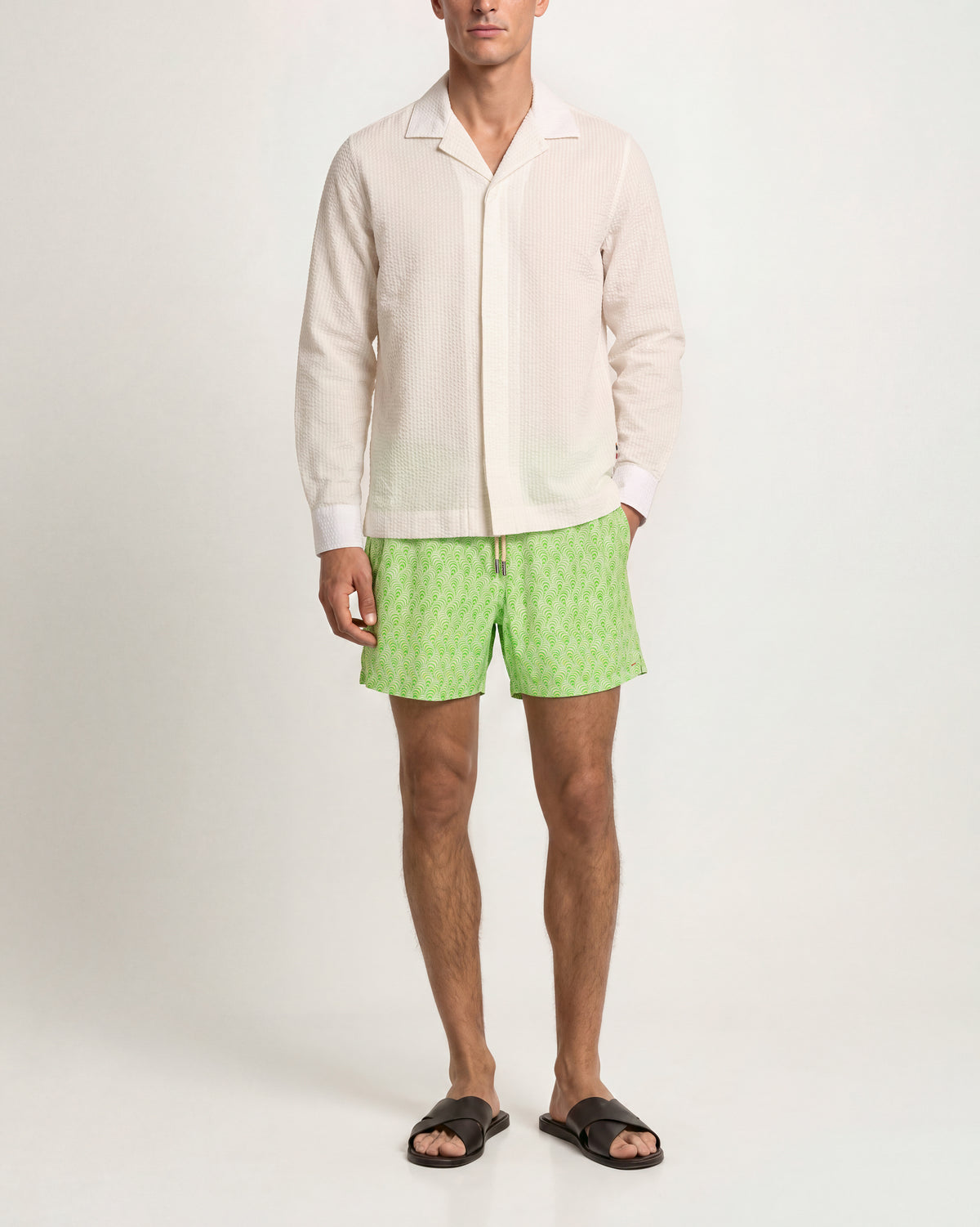 A male model wearing a cream-colored long-sleeve camp collar shirt and light green patterned swim trunks, paired with black slide sandals, against a plain light gray studio background.