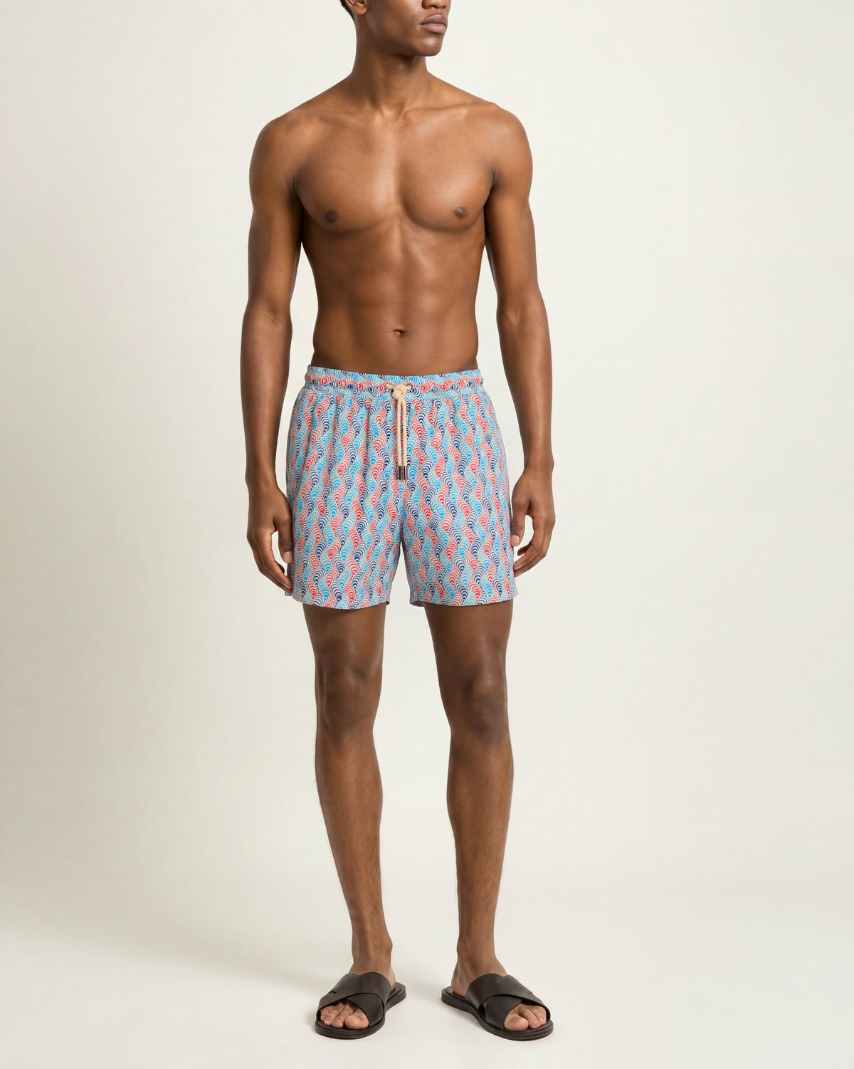Man wearing blue and pink patterned swim shorts with brown sandals on a plain background