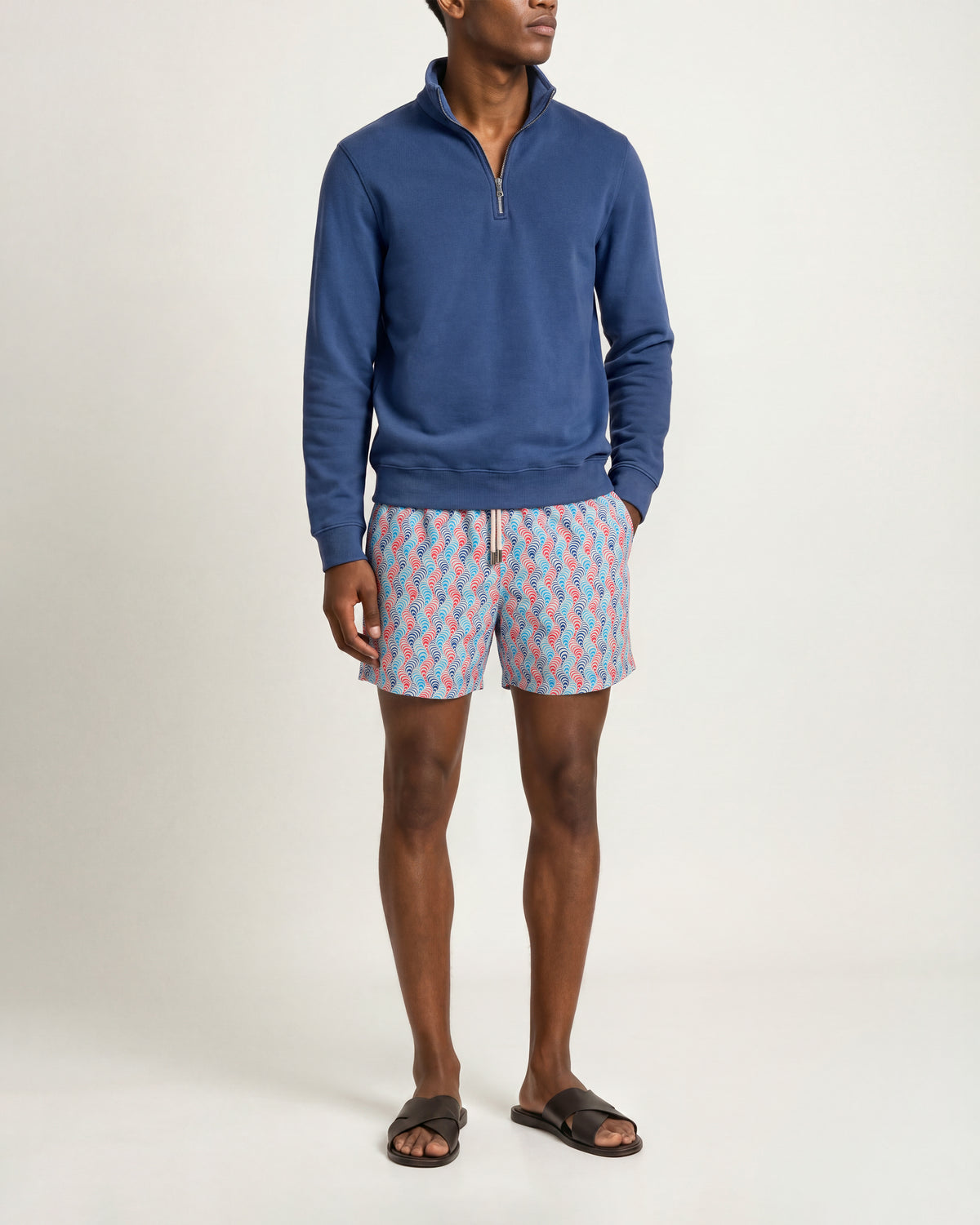 A man modeling a blue long-sleeve quarter-zip pullover and patterned shorts with brown slide sandals, photographed against a plain light gray studio background.