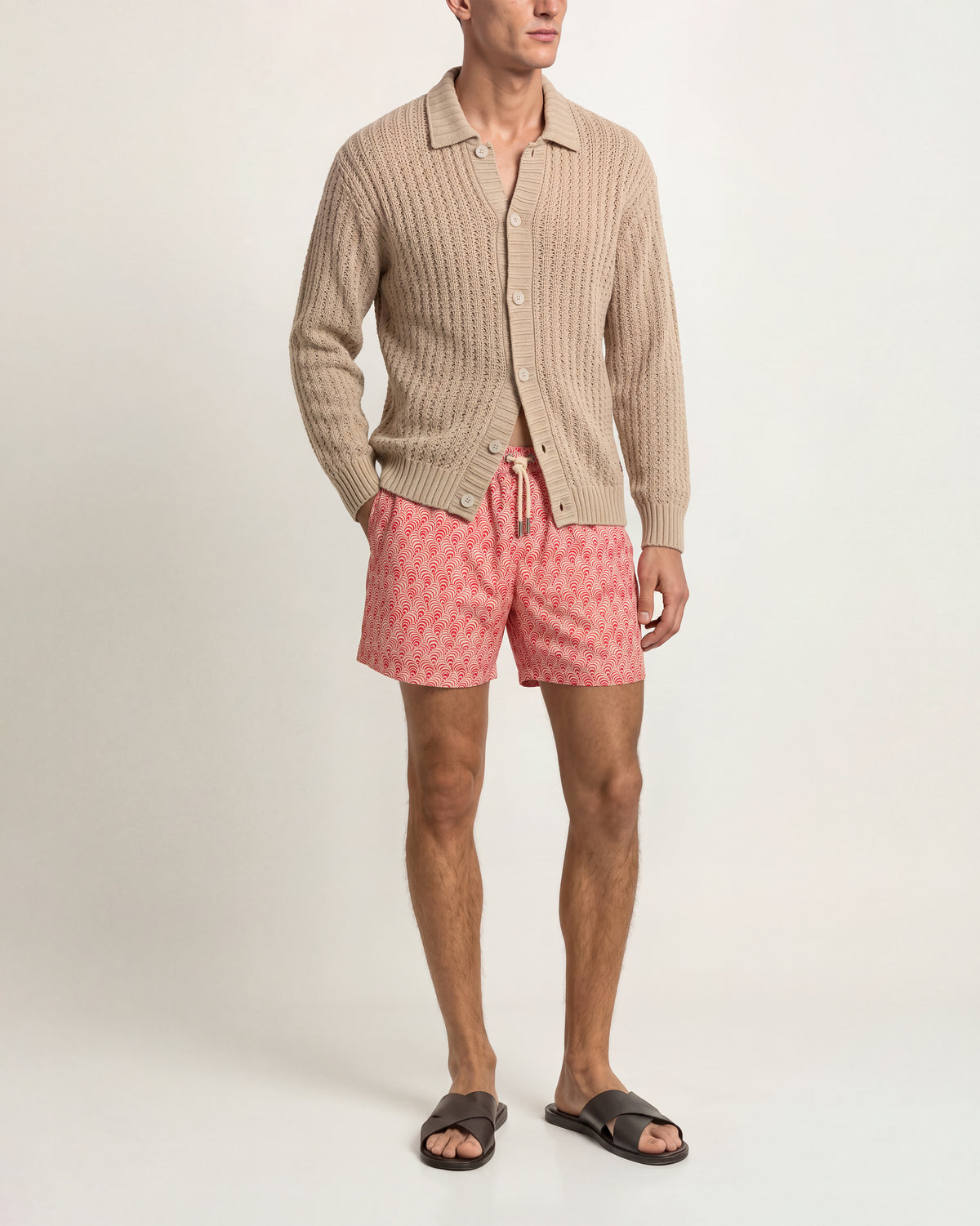A man modeling a beige cable-knit button-up cardigan, pink patterned shorts, and dark brown slide sandals against a plain light background.