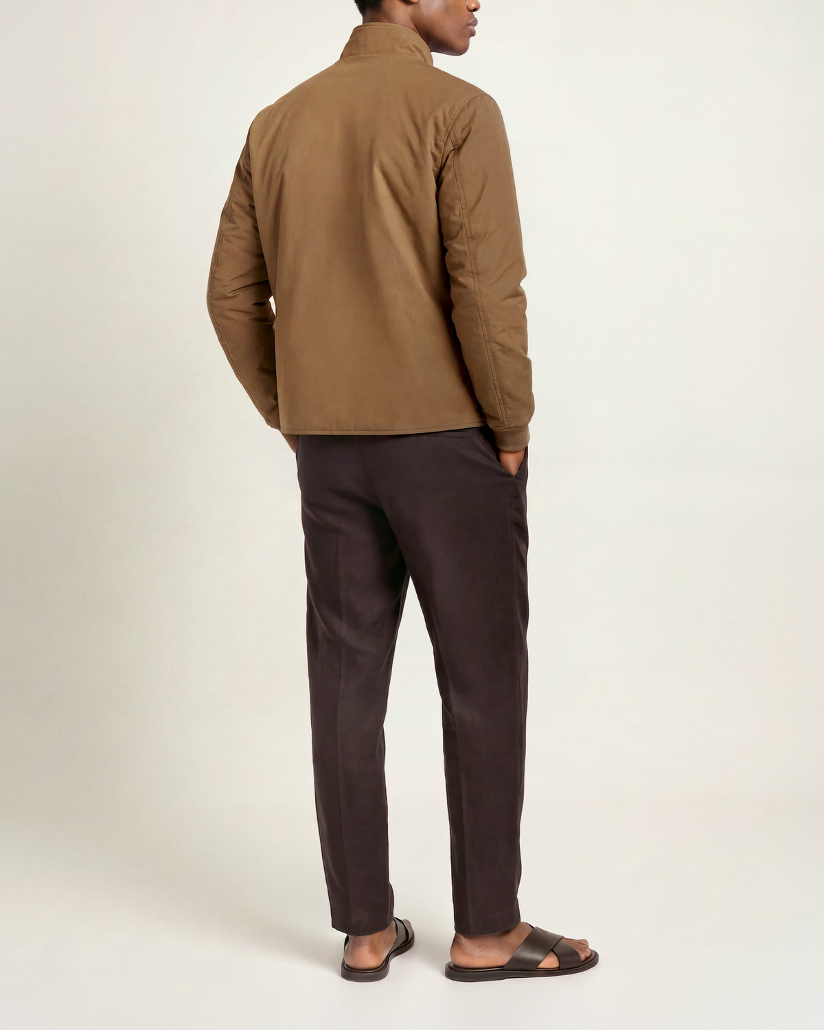 Man wearing bronze coloured padded jacket with brown linen trousers and sandals on a plain background