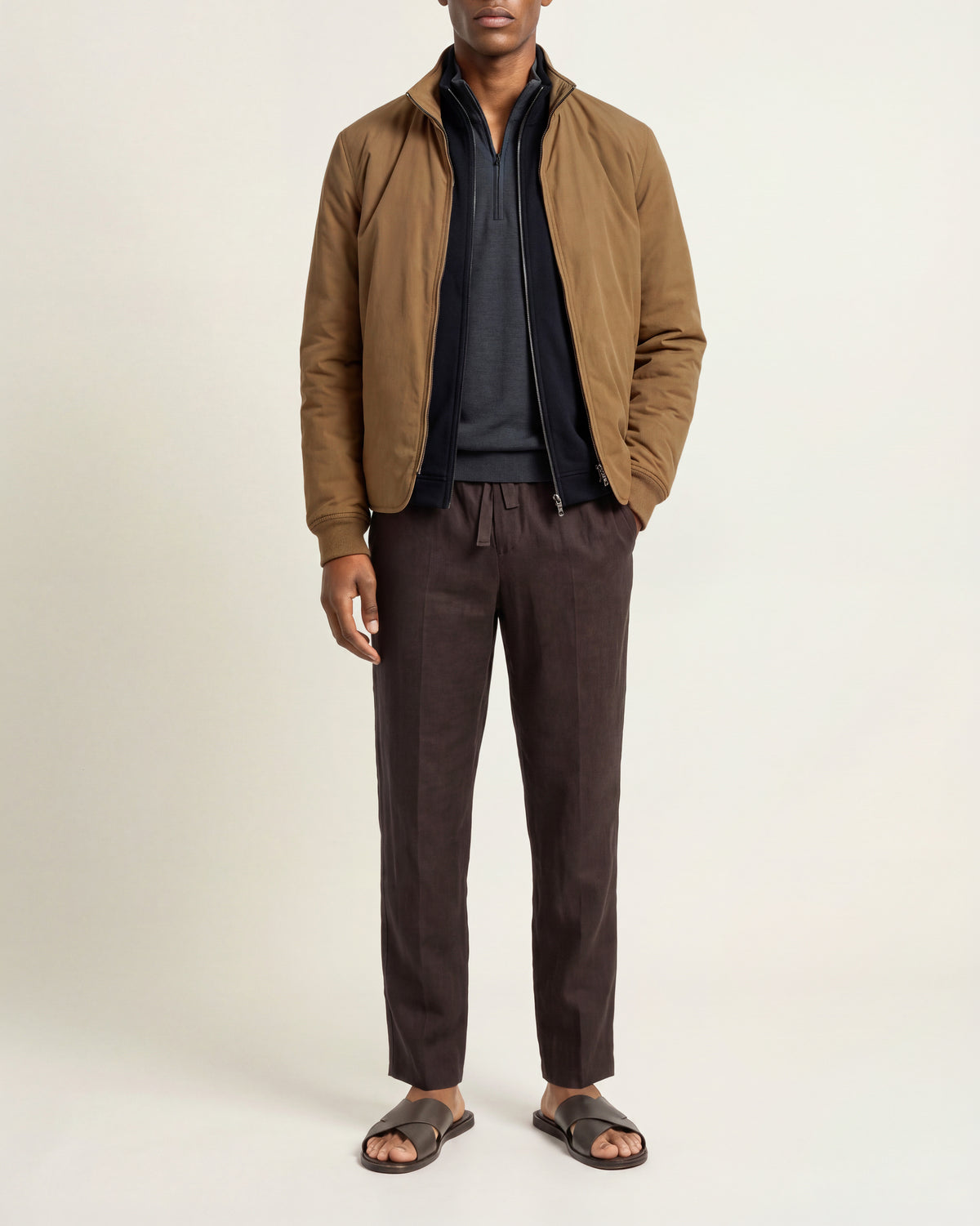 Man wearing bronze coloured padded jacket with navy and grey zip-up underneath. Worn with brown trousers and sandals on a plain background