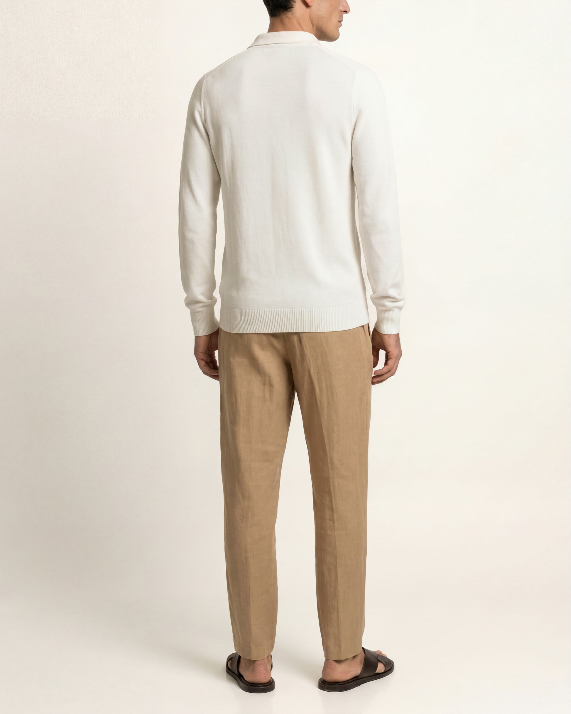 A man shown from the back wearing a cream long-sleeve knit polo shirt and beige trousers, standing against a plain off-white studio background.