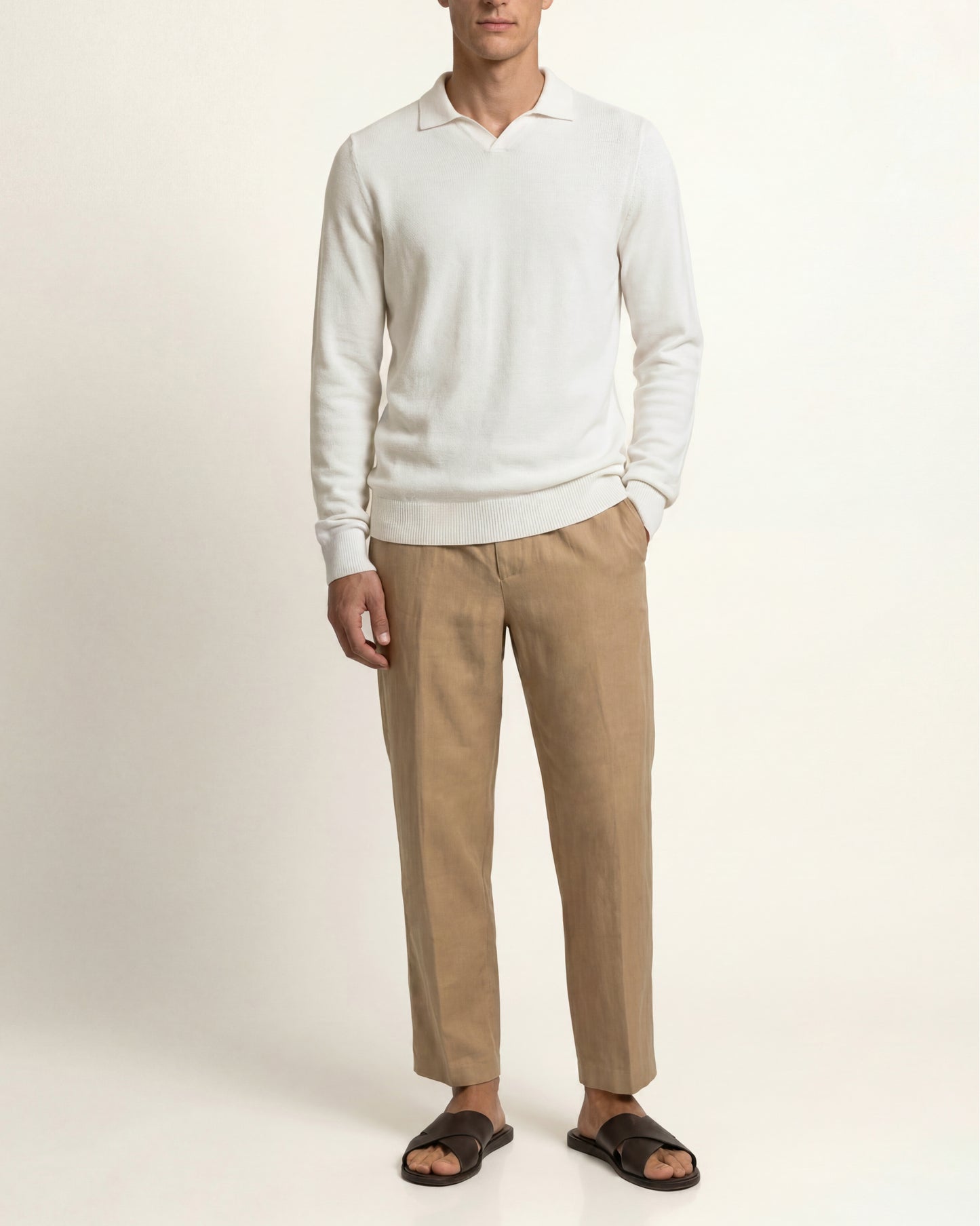 A man wearing a white long-sleeve polo sweater, beige trousers, and dark brown slide sandals, standing against a plain off-white studio background.