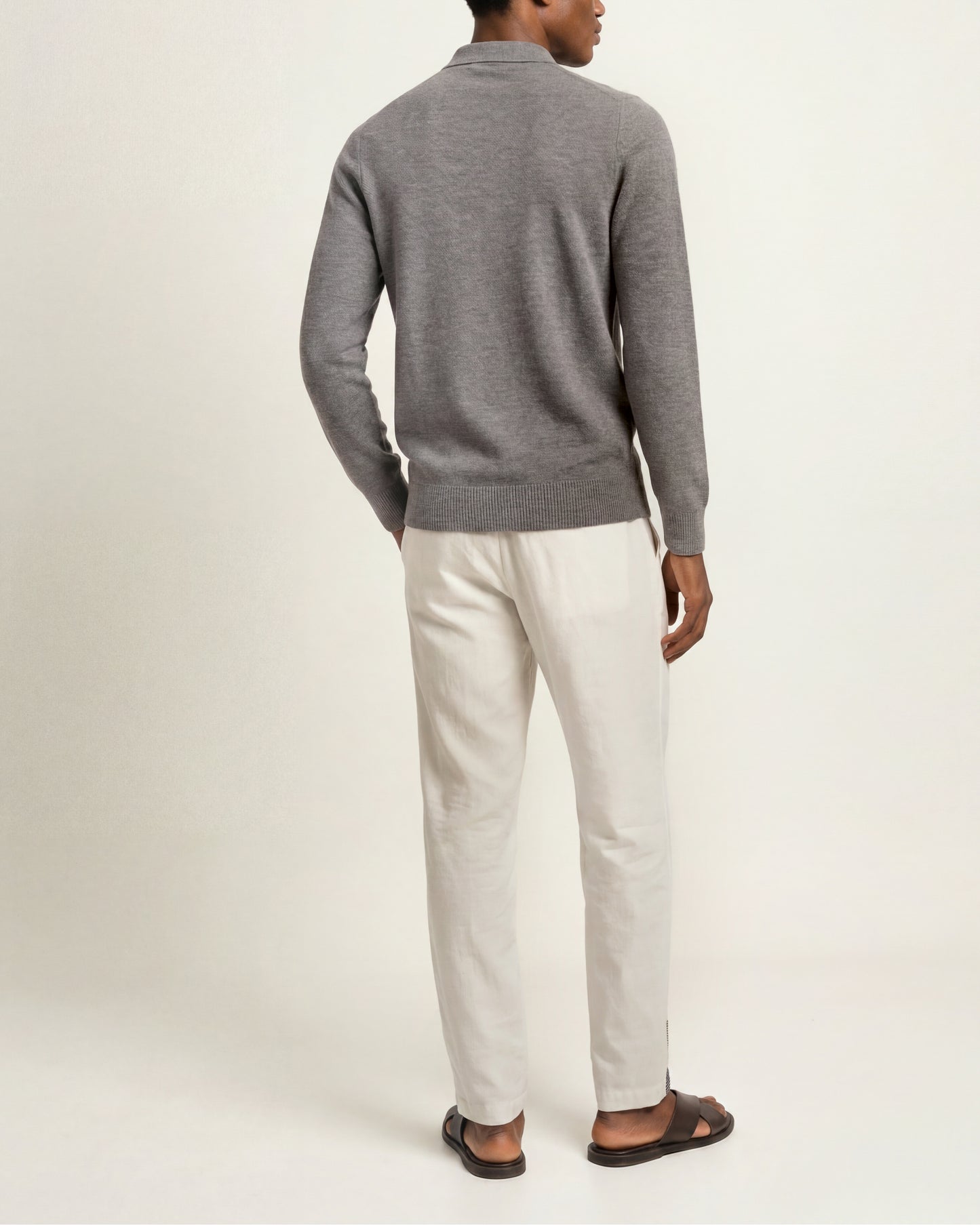 A man shown from the back wearing a gray knit quarter-zip sweater, off-white trousers, and dark brown sandals, standing against a plain light beige studio background.