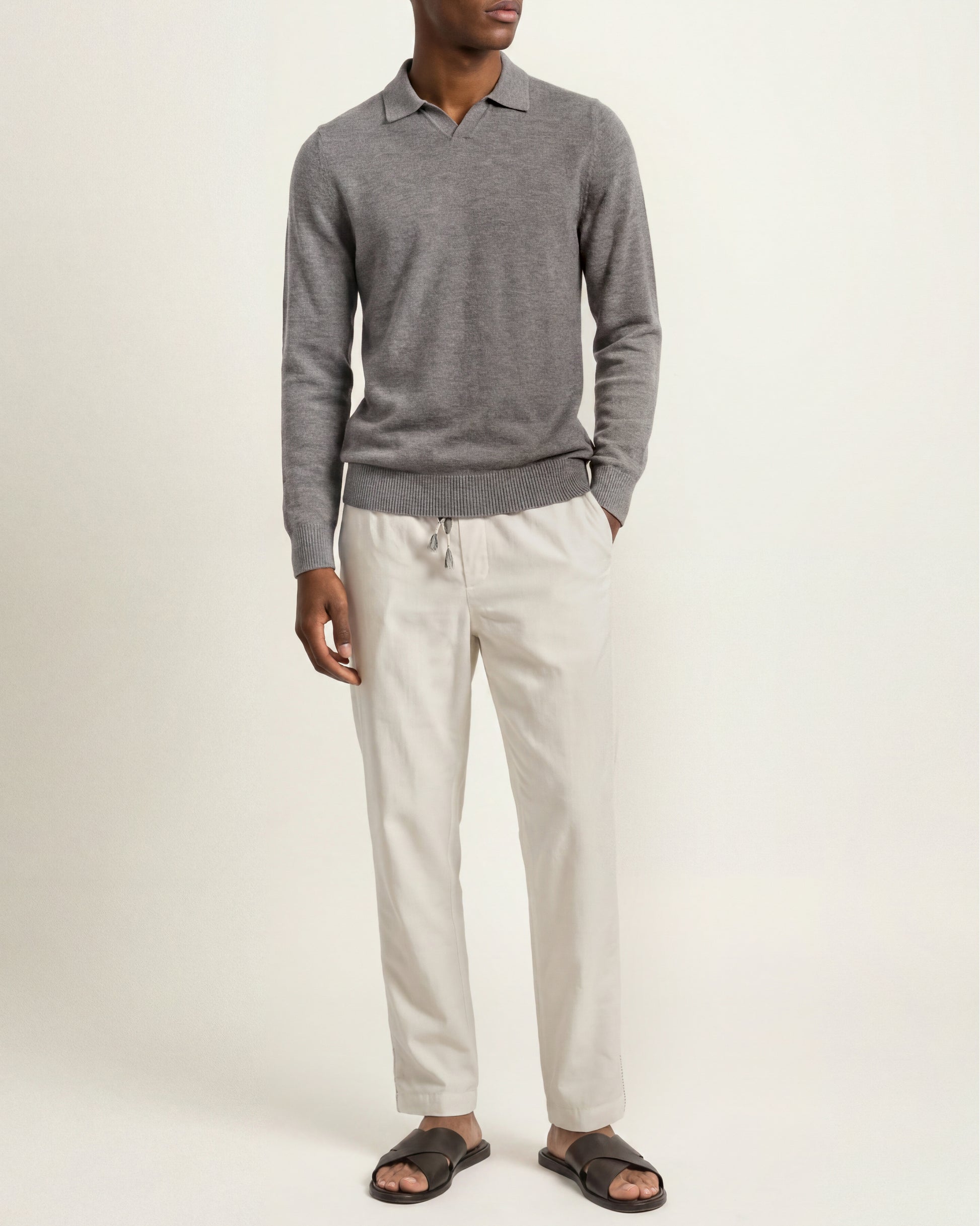 A man wearing a gray long-sleeve polo sweater, off-white trousers, and dark brown slide sandals, standing against a plain light beige studio background.