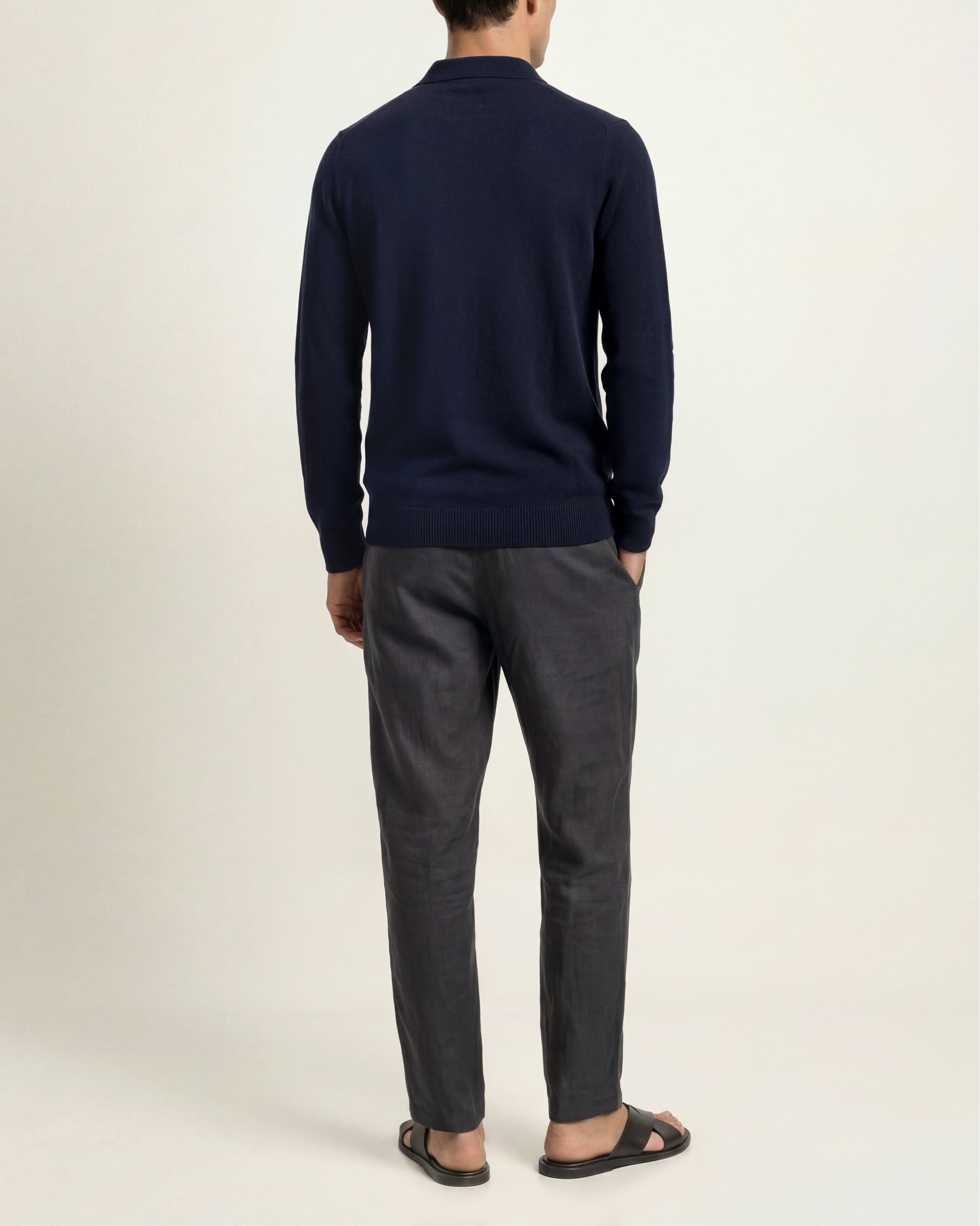 A man viewed from the back wearing a navy blue long-sleeve knit sweater, dark charcoal gray trousers, and black slide sandals, standing against a plain off-white studio background.