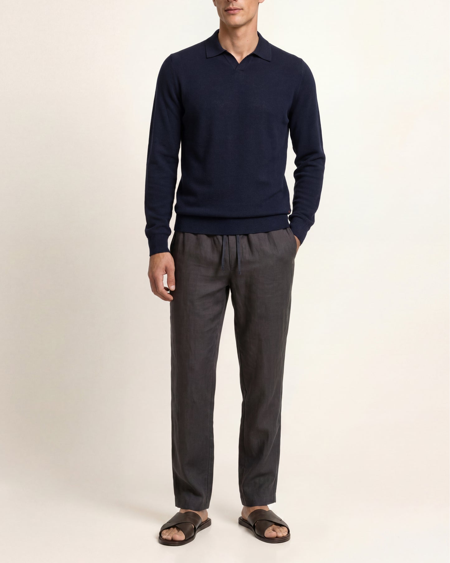 A man modeling a navy blue long-sleeve polo sweater paired with dark gray drawstring trousers and dark brown slide sandals, photographed against a plain off-white studio background.