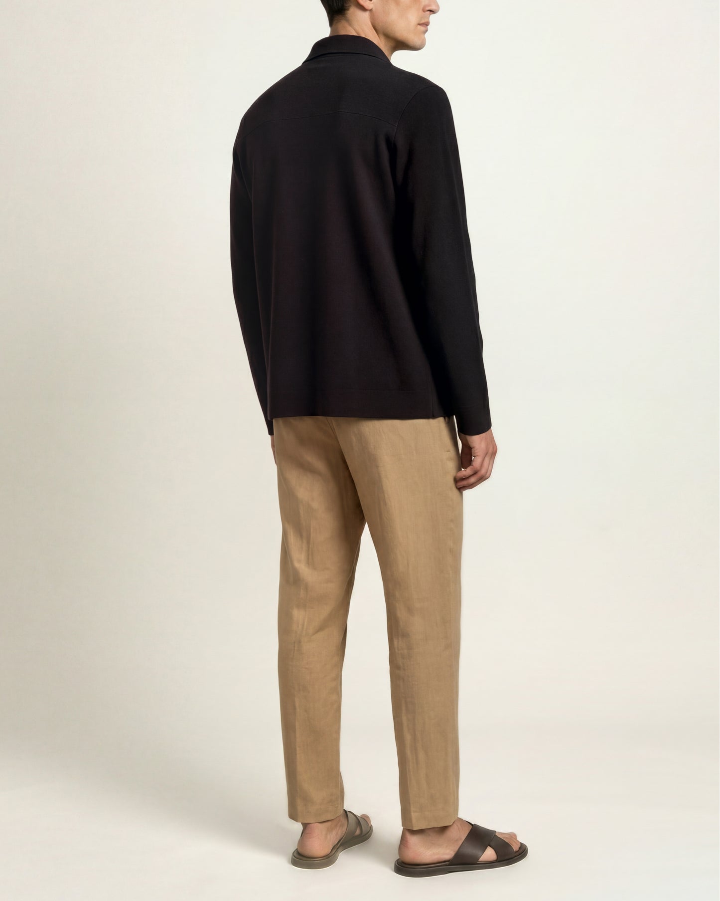 A man shown from the back wearing a black long-sleeve knit top, beige trousers, and dark brown sandals against a plain off-white studio background.