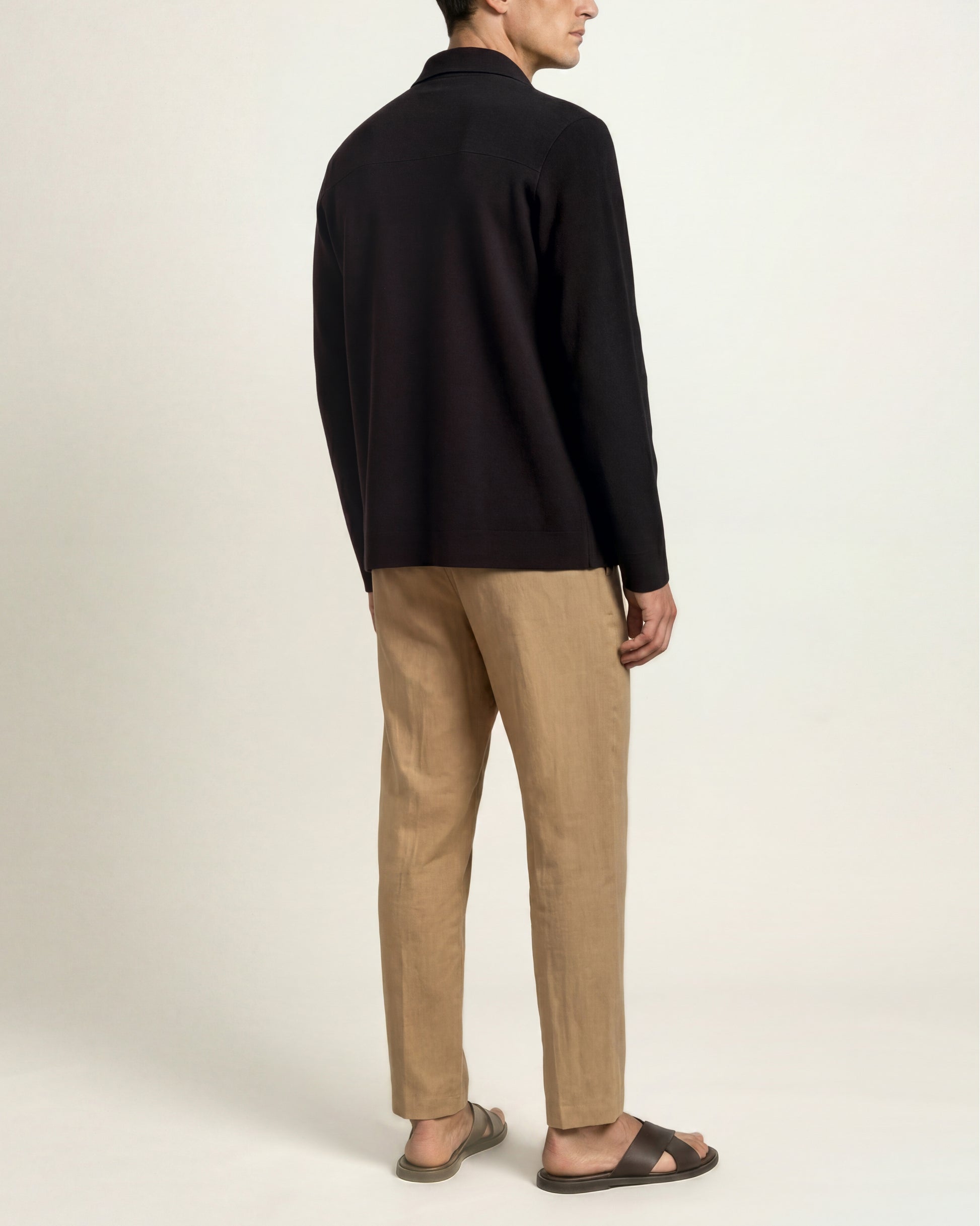 A man shown from the back wearing a black long-sleeve knit top, beige trousers, and dark brown sandals against a plain off-white studio background.