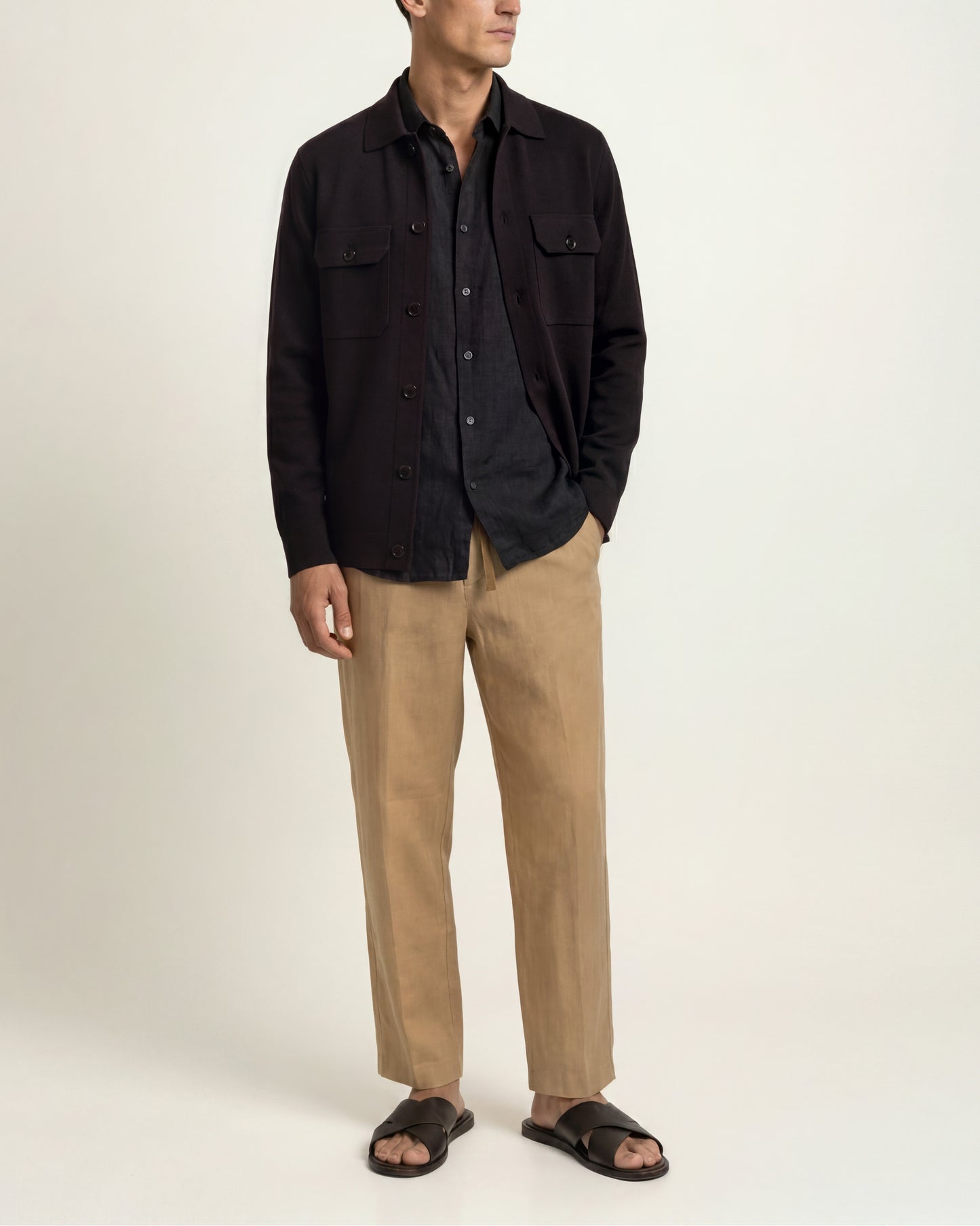 A man modeling a black button-up overshirt over a dark shirt, paired with beige trousers and black slide sandals, against a plain light background.