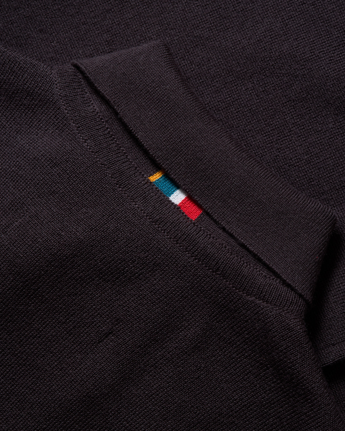 Close-up of a dark charcoal knit polo shirt collar featuring a small embroidered multicolor stripe in yellow, teal, white, and red.