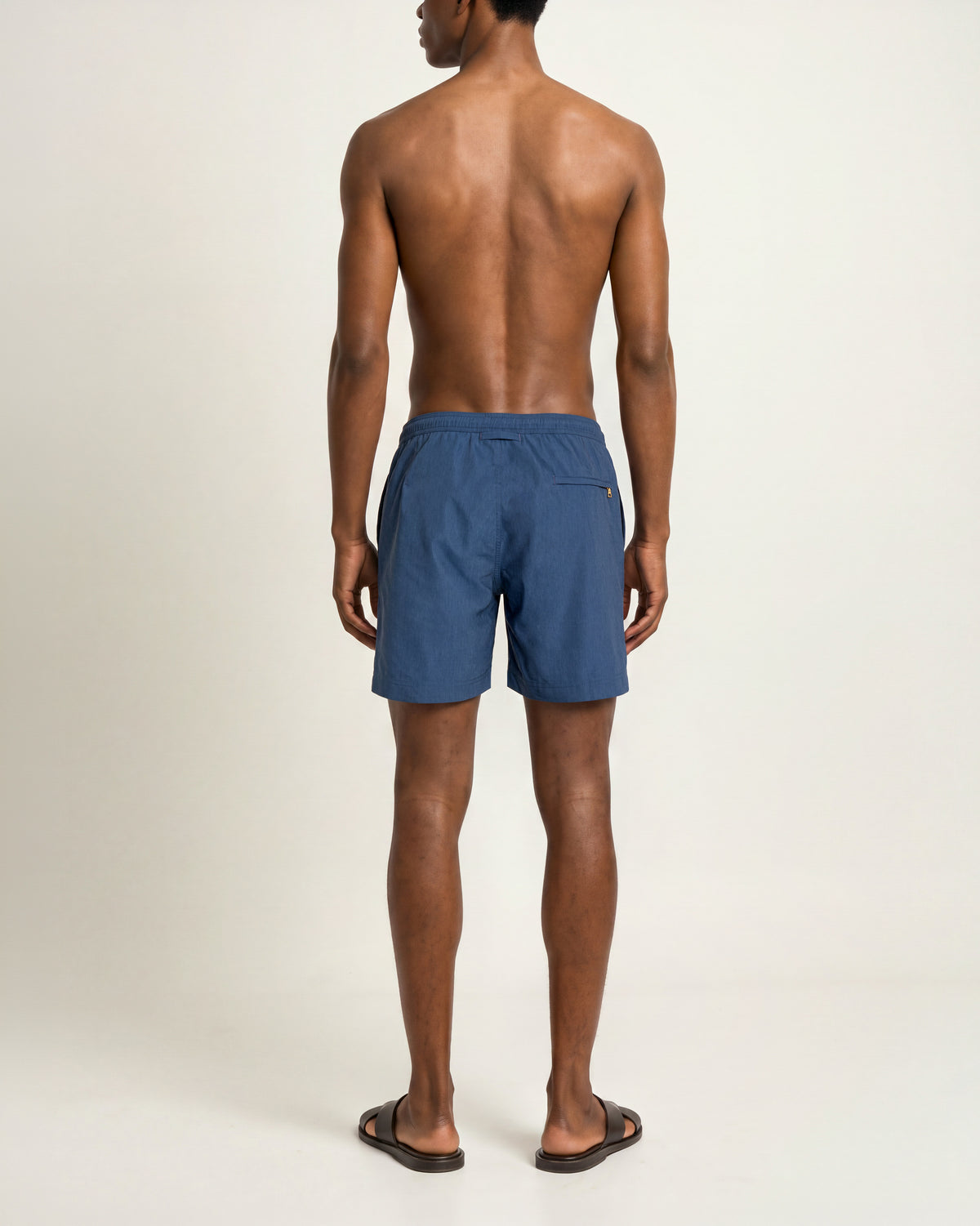 Back view of a man wearing aquamarine drawcord swim shorts and brown sandals on a plain background