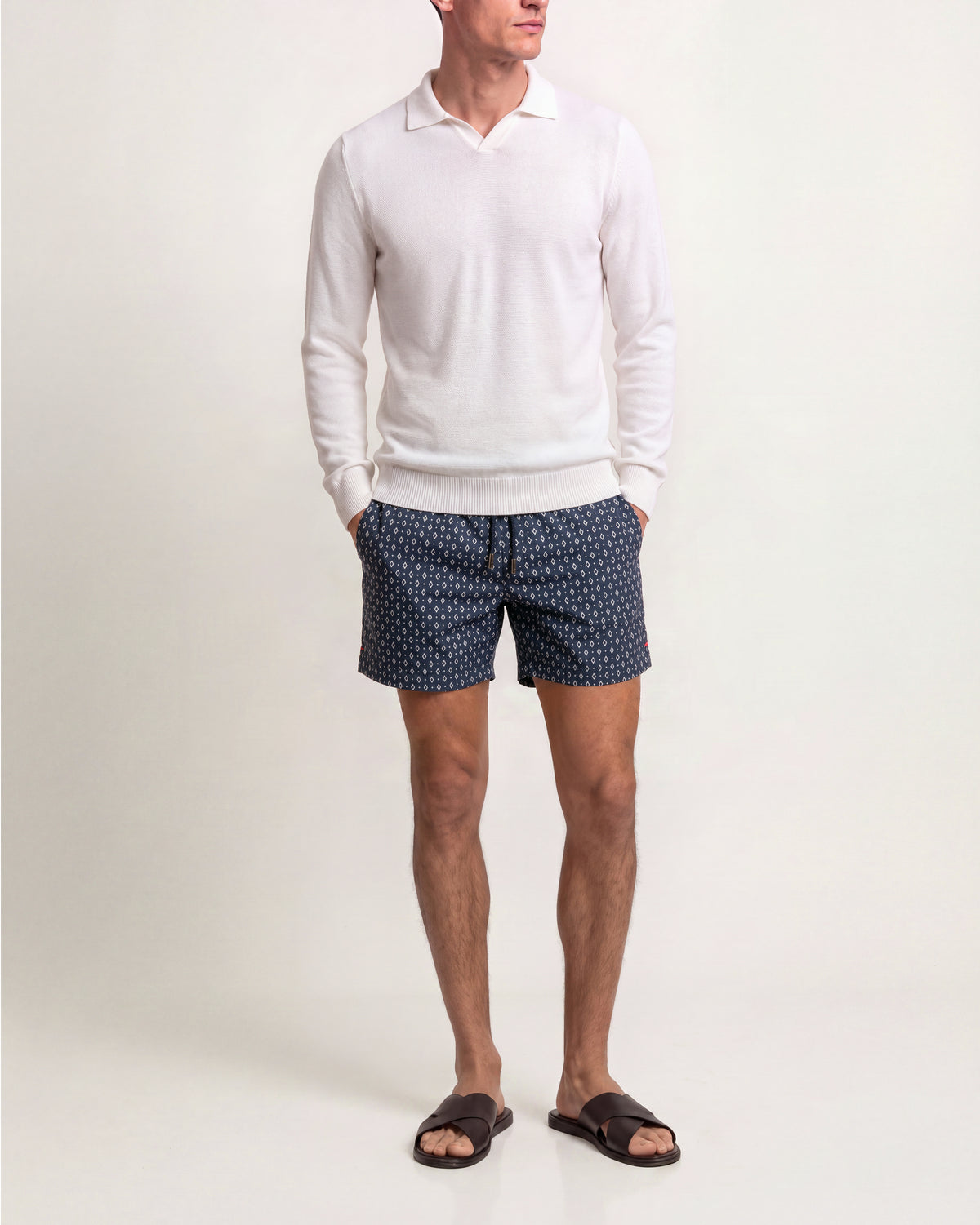 A man wearing a white long-sleeve polo sweater, navy blue shorts with a small white geometric pattern, and dark brown slide sandals, standing against a plain off-white studio background.